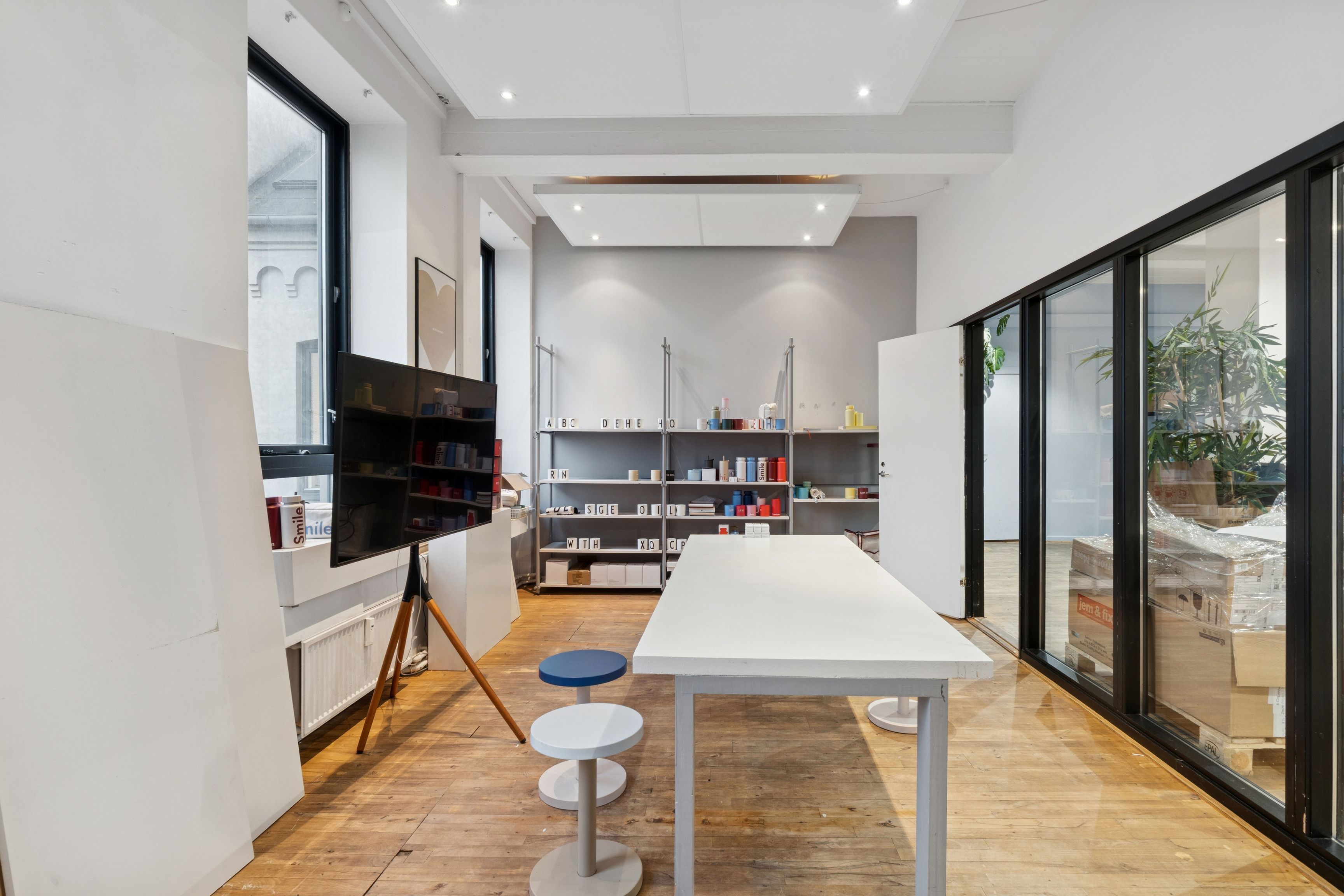 En stor rummelig kontor med hvid væg, høj loft og trægulv, indeholder flere møbler, herunder høj bord, stole og et højt stativ med tv