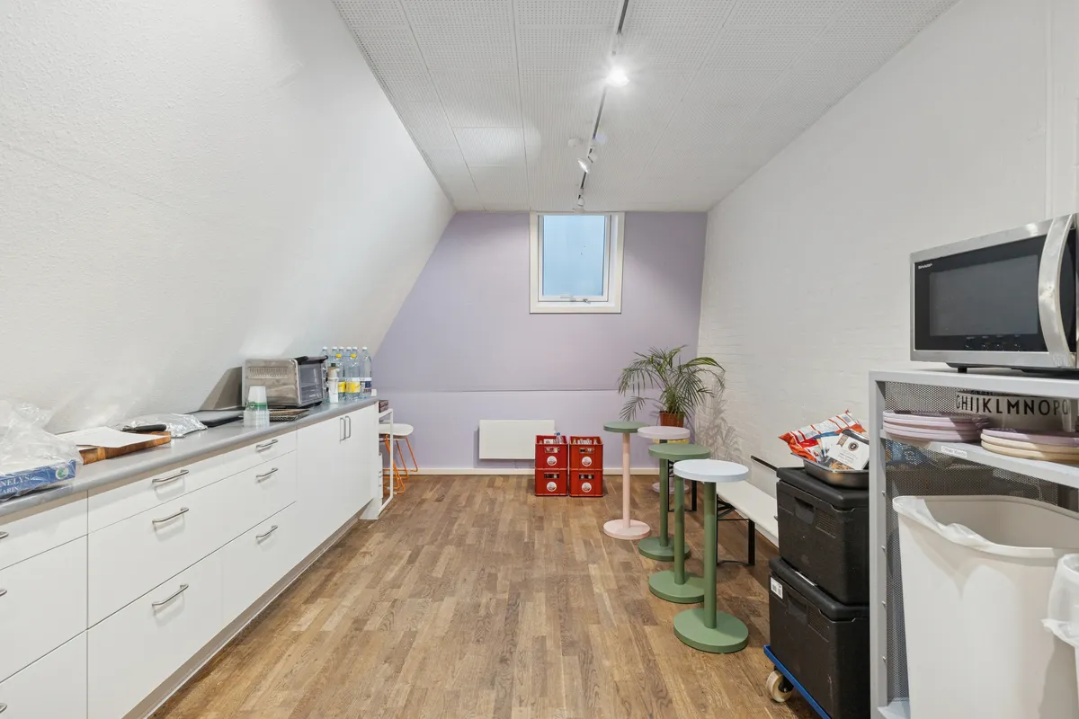 Storrumskontor med loftsrum, højloft, to mikrobølgeovne, to høje stole, plastbokse og brædder, to lyse stolpelampe og trægulv