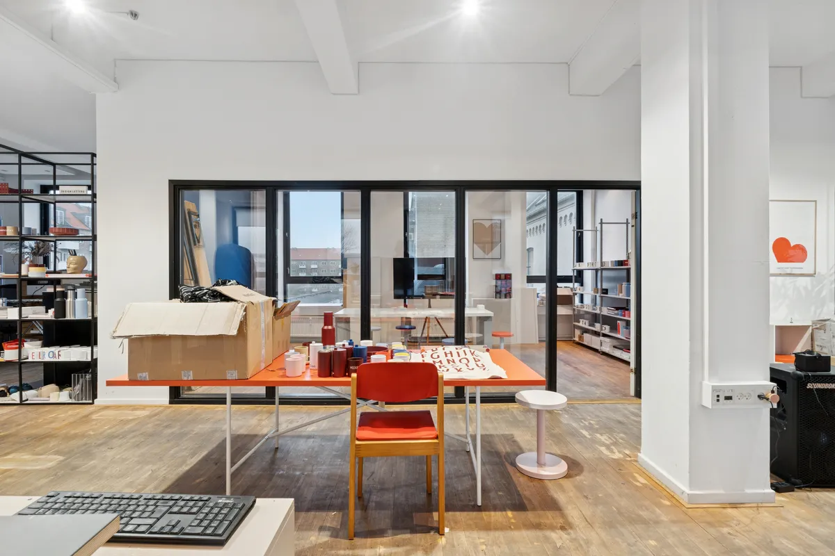 Inde i et storrumskontor med glasvinduer og loftsbjælker er der en stor orange bord med en række farvede krus og skriftstykker samt en åben boks