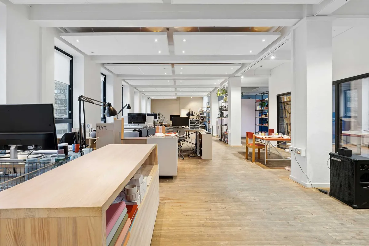 Moderne kontor med lyse lofter og hårde trægulve. Flere borde med computere og møbler samt en lysebrun sofa. Styret lys skaber en velkomststemning i kontoret.