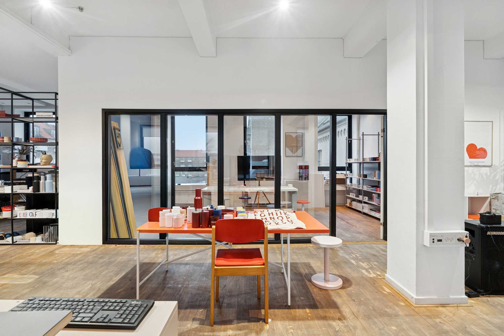 Moderne kontor med glasvægge, orange bord, højstole, keyboard og små stole. Det er et åbent rum med masser af lys, og det er delt i to områder med en stolpe i midten.