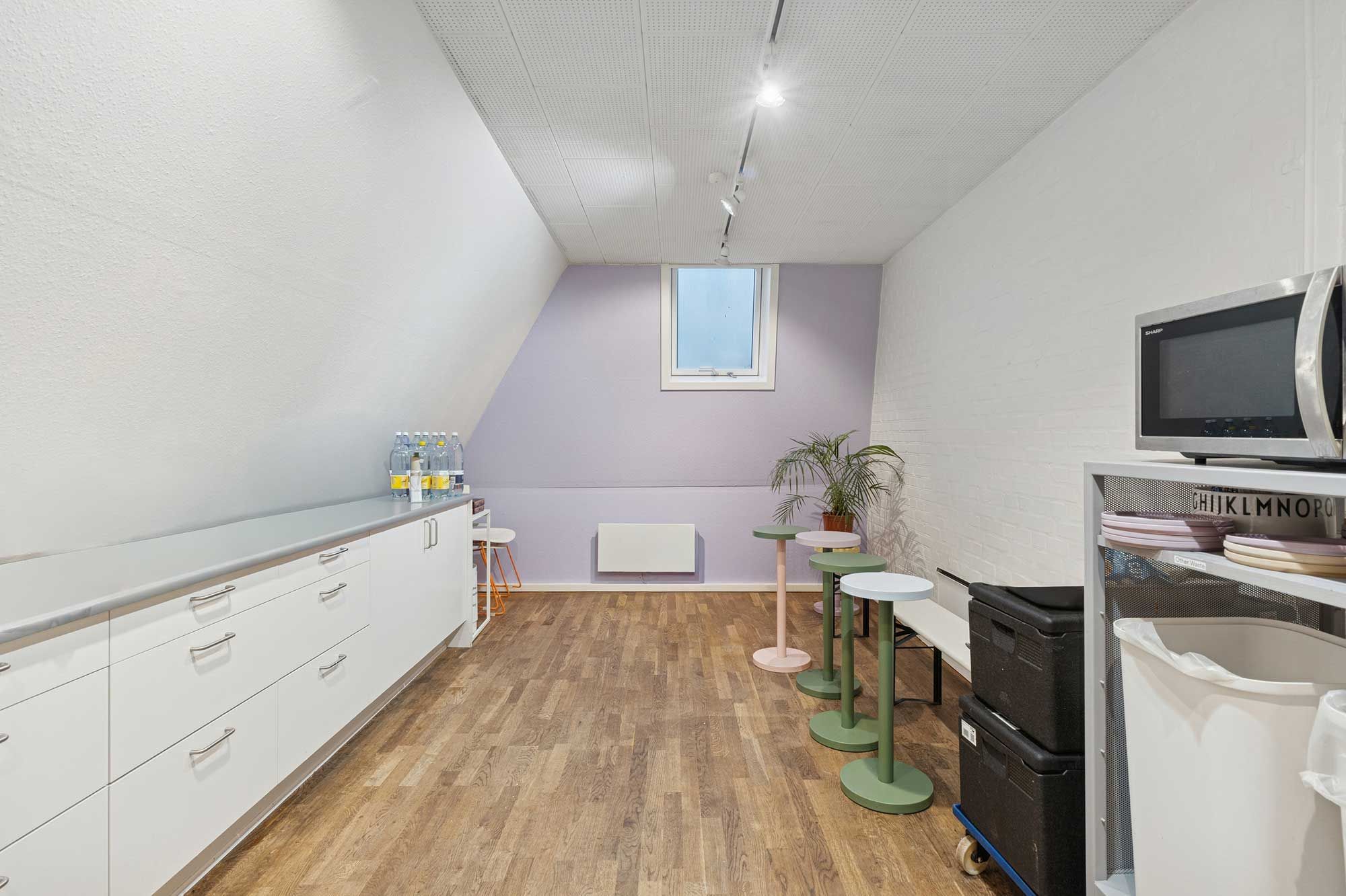 Rummeligt kontor med moderne møbler og højloft. Vindue, mikroovn, lysekroner og grønne planter. Gulvbelægning og lysegrå vægge.