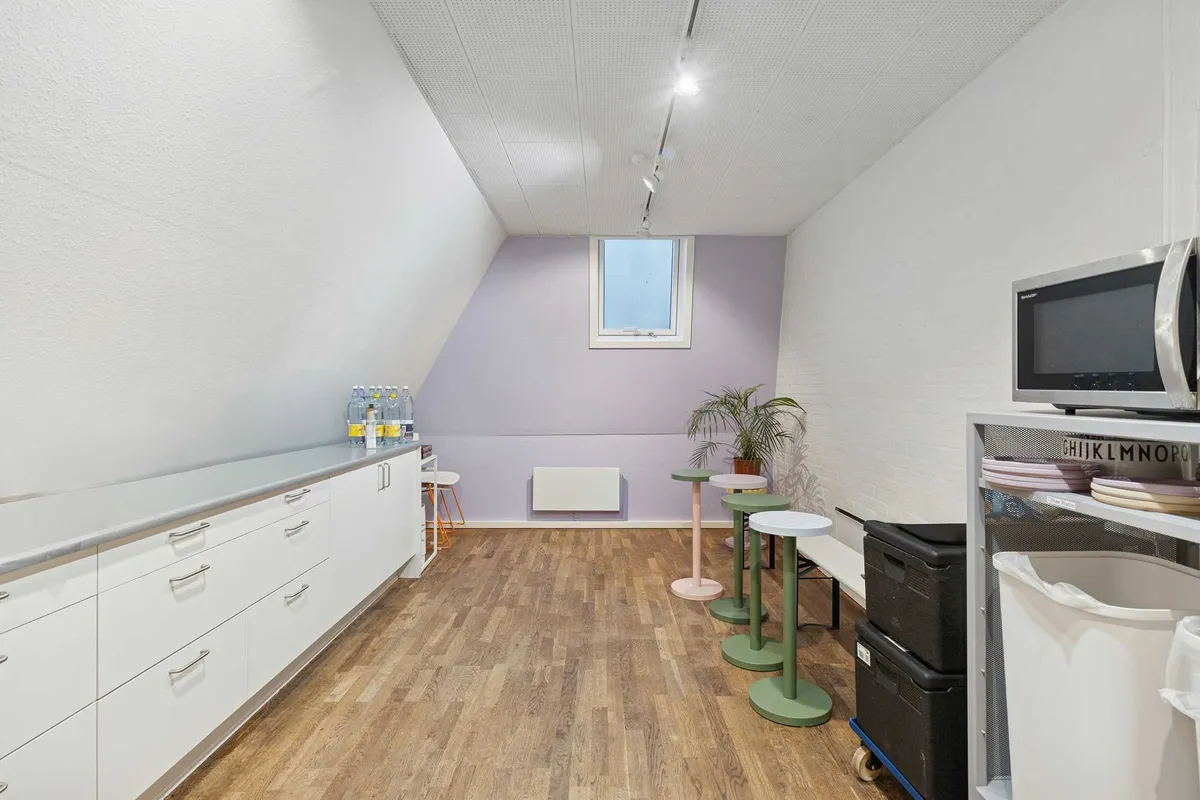 Rummeligt kontor med moderne møbler og højloft. Vindue, mikroovn, lysekroner og grønne planter. Gulvbelægning og lysegrå vægge.