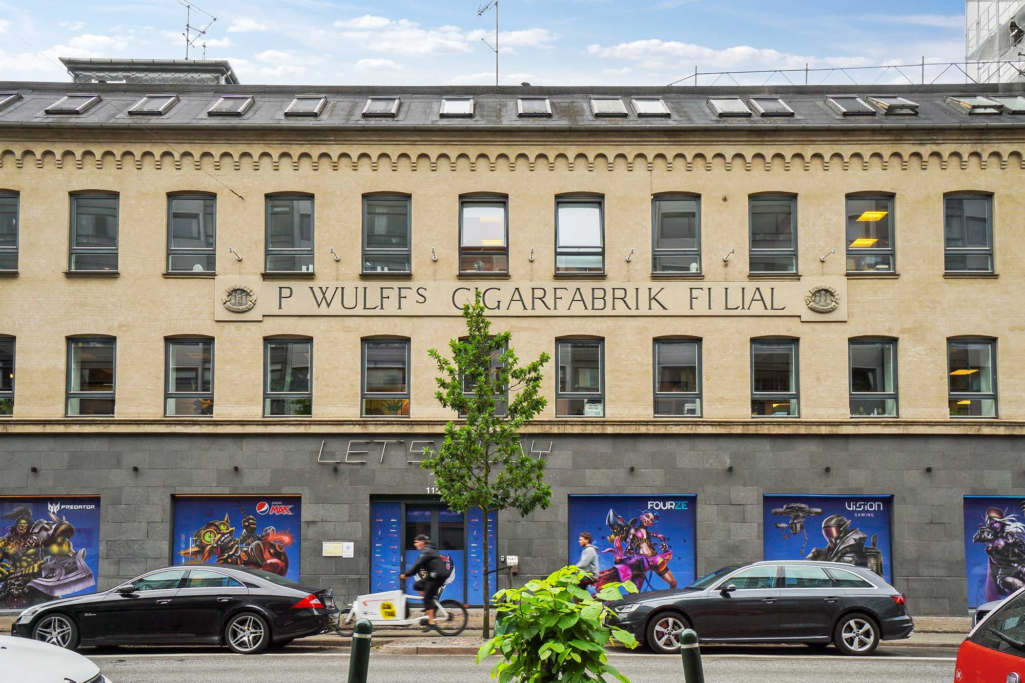 Et stort kontorbyggeri i P. Wulffs Cigarfabrik i København med moderne reklamebanner på facaden og biler parkeret foran