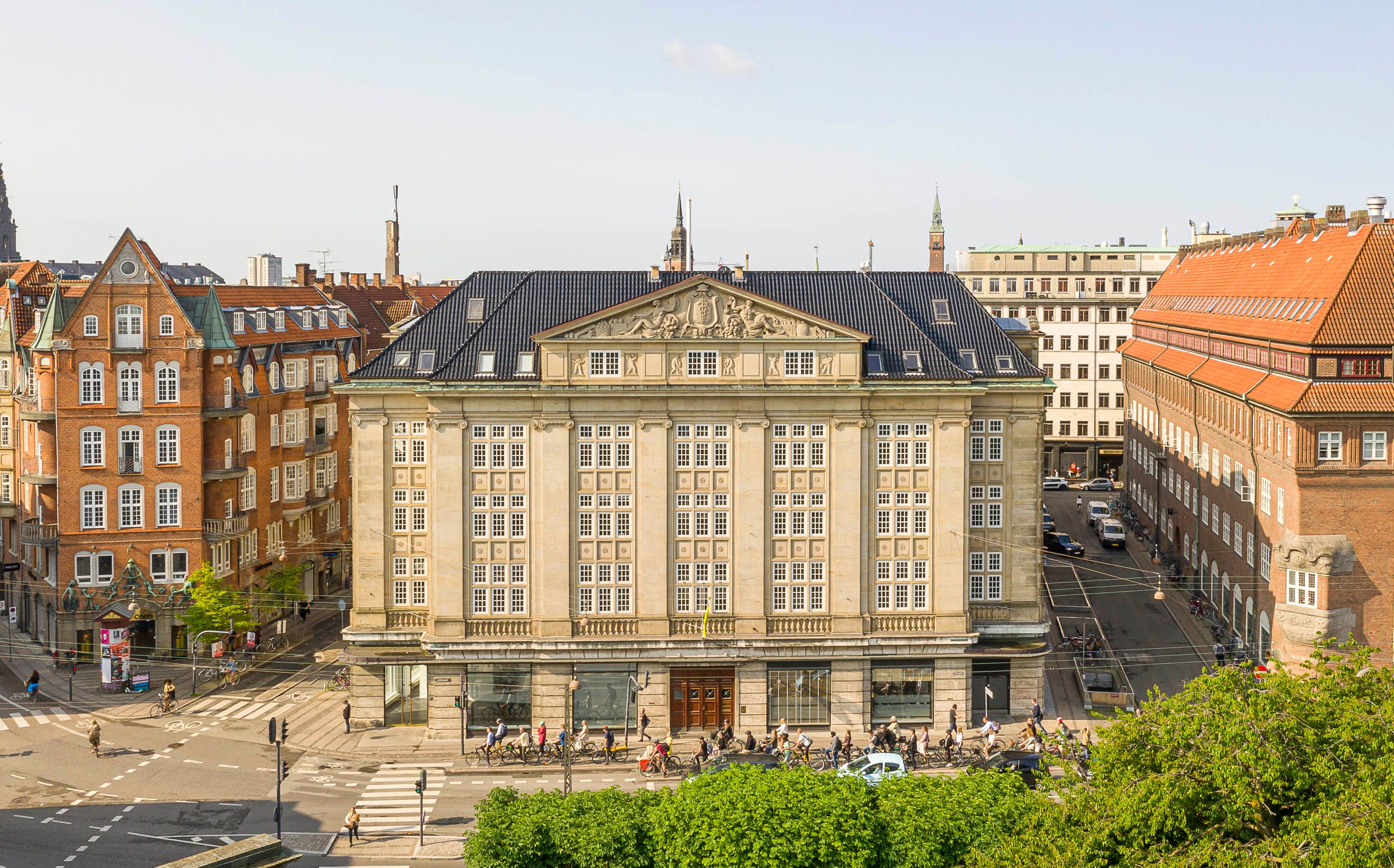 Københavns centrum med mange mennesker, der cykler og går på gågaden. Bygninger med røde tag og mange vinduer og træer