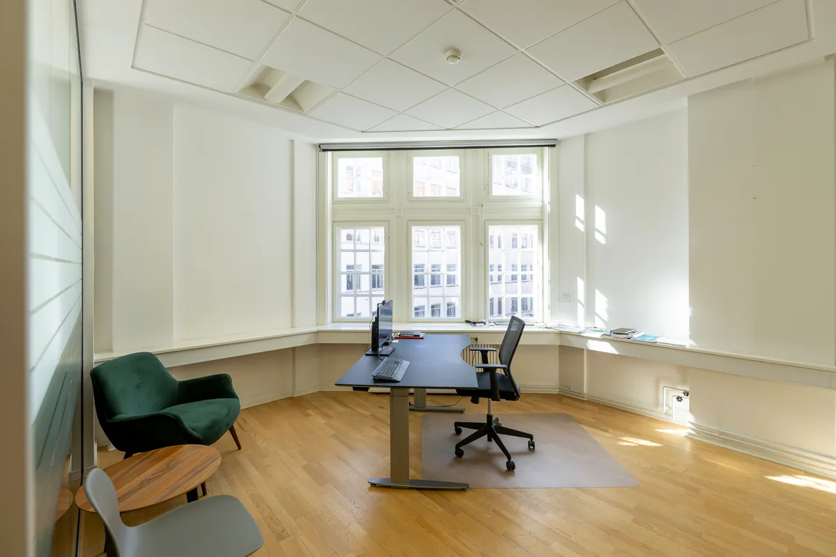 Luksuriøst kontor med højt gulv og moderne møbler. Et skrivebord med en computer og en stol står midt i rummet. Væggene er hvide, og vinduer med gardiner giver masser af lys.