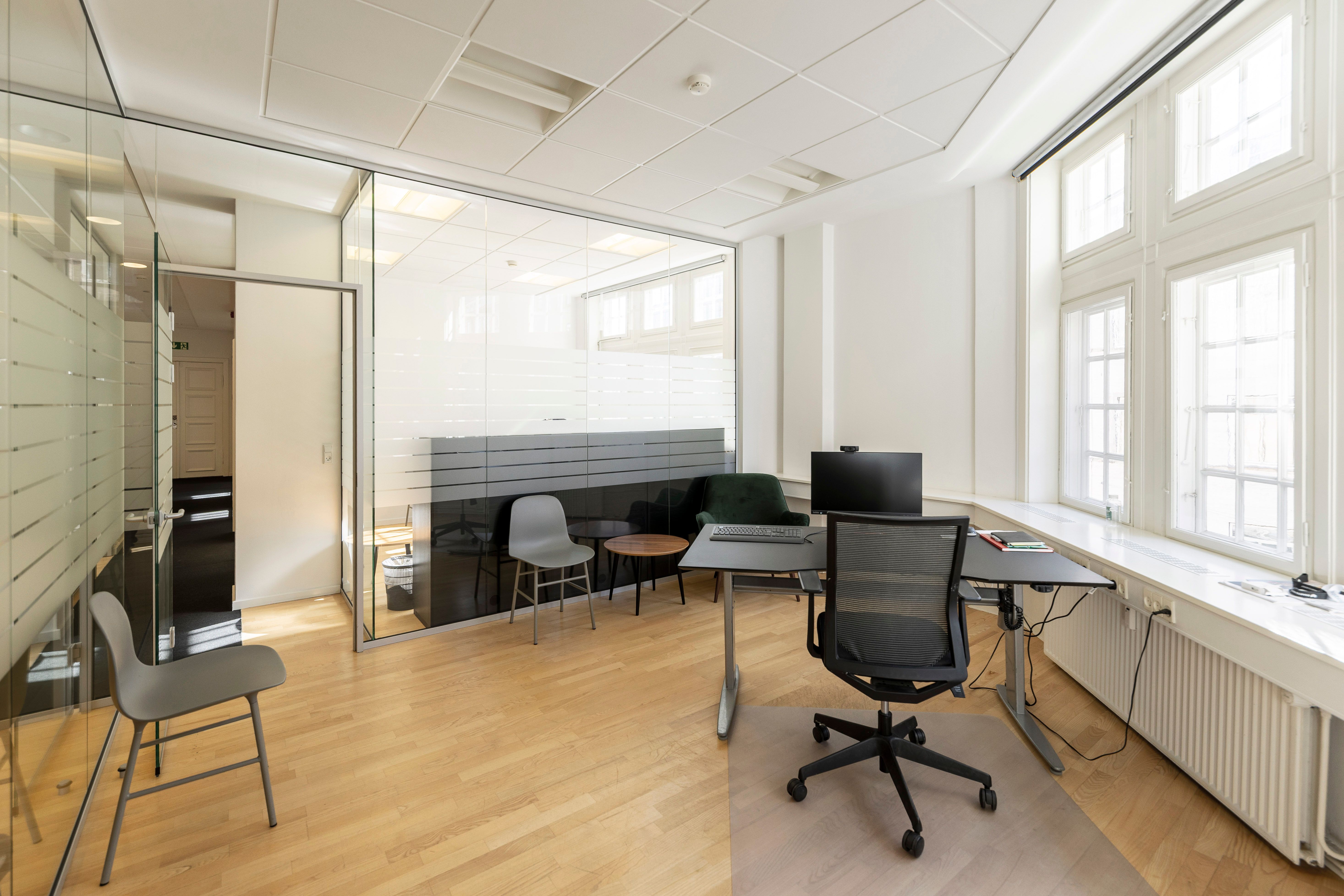 Kontor med moderne indretning, lyse vinduer, højloft, trægulv, to skriveborde, to stole og radiatorer.