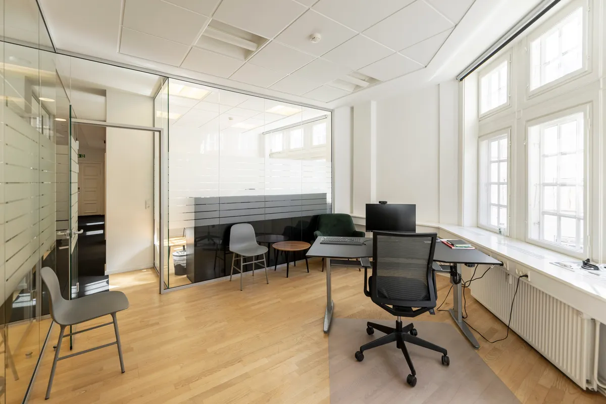 Kontor med moderne indretning, lyse vinduer, højloft, trægulv, to skriveborde, to stole og radiatorer.