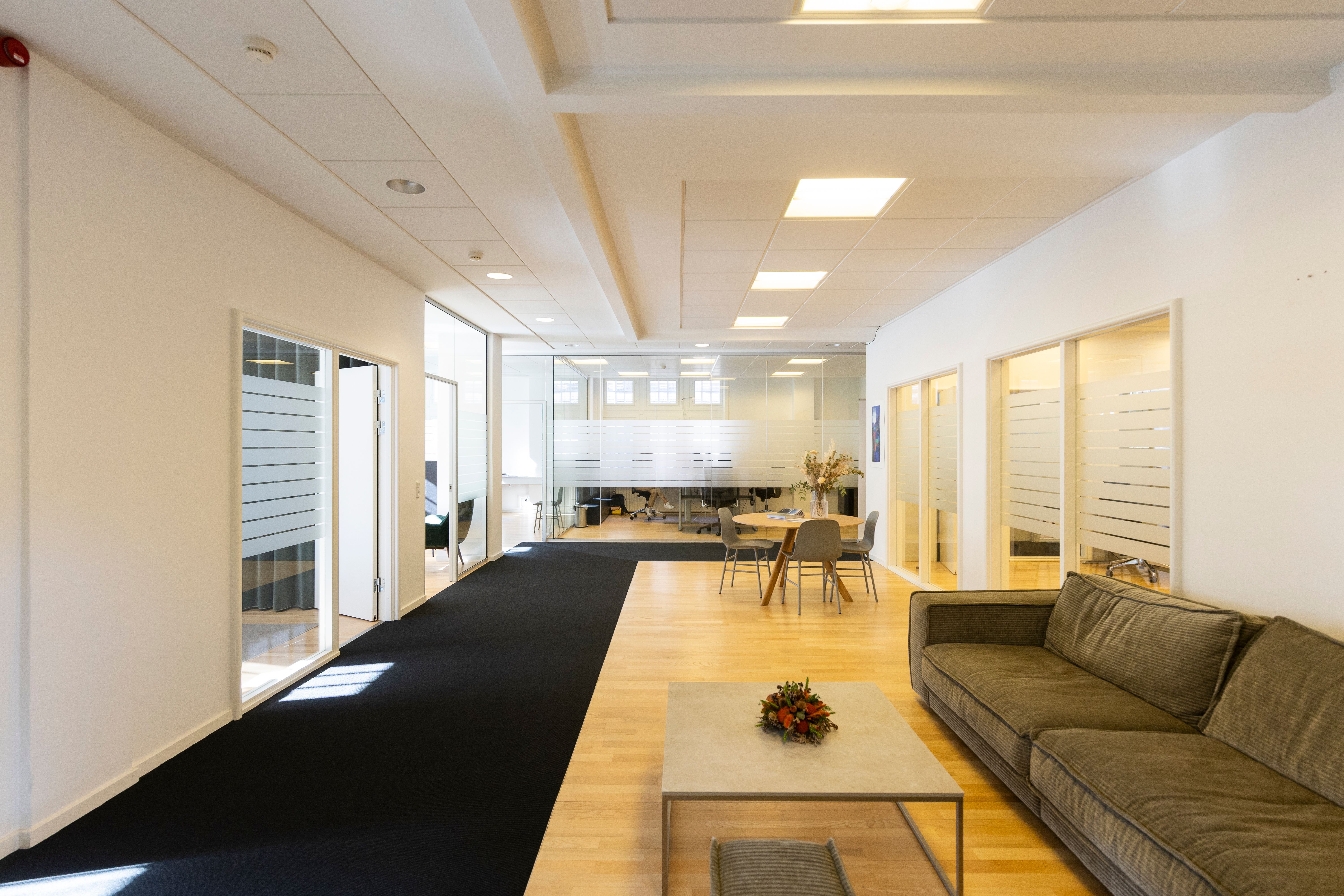 Indre af kontor med sort gulvtæppe, møbler og lyse lofter samt store glasvinduer, der vender ud mod en fælles kantine og udendørsarealer