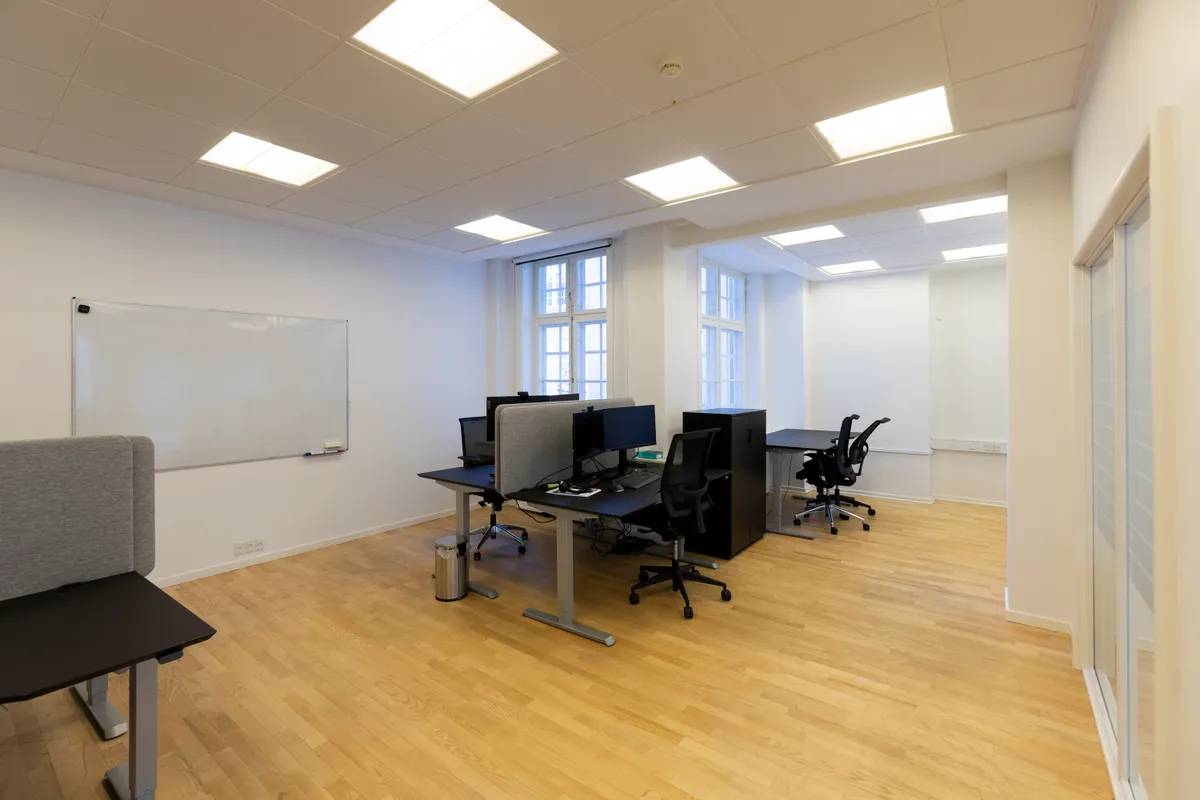 Moderne kontorrum med lyse loftslampe, høje ruder, lysegult gulv og minimalistisk møblering inklusive borde og skriveborde