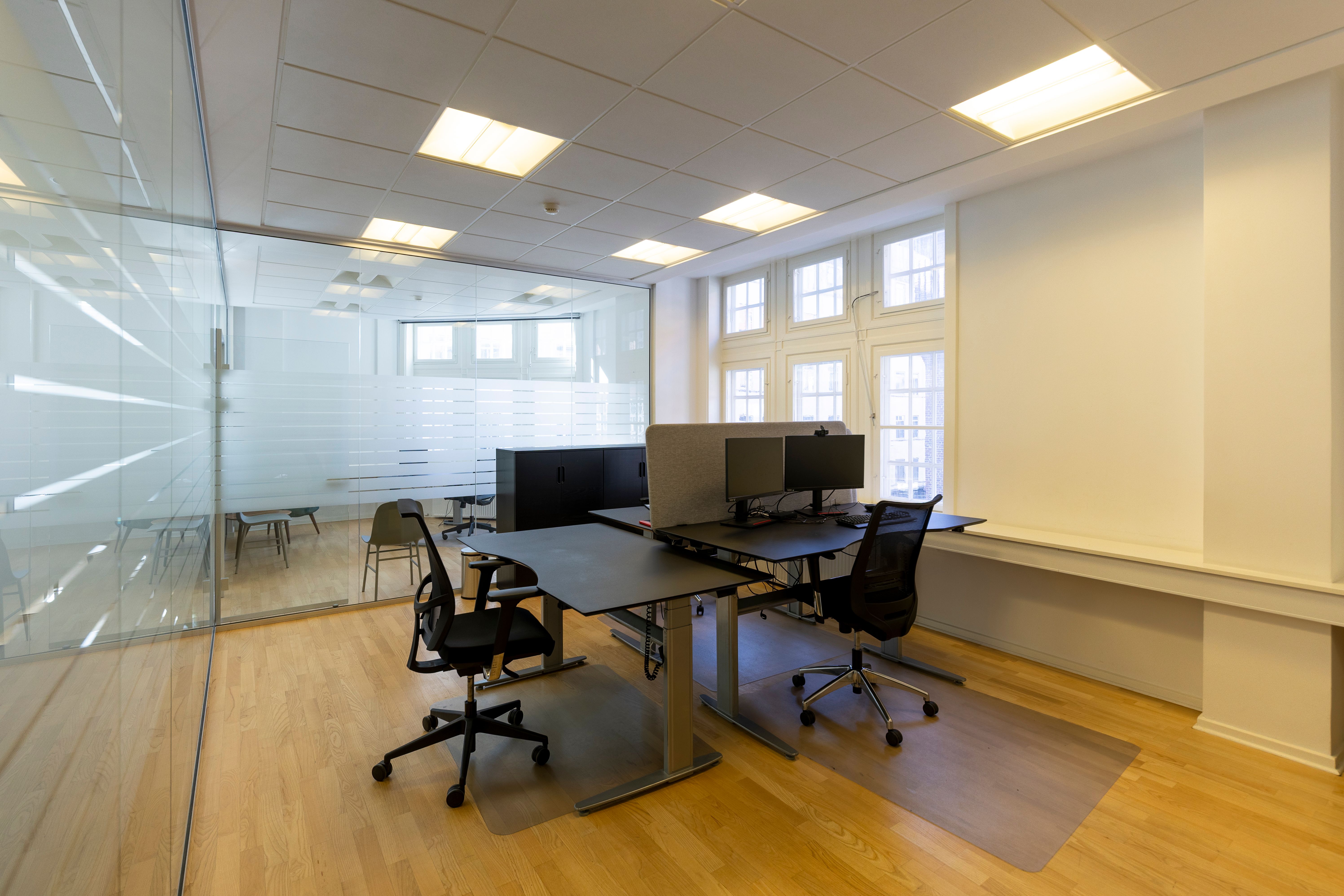 Kontorrum med store vinduer og moderne møbler samt højt loft. To bord med skrivebord og stol samt en partition og kantiner.