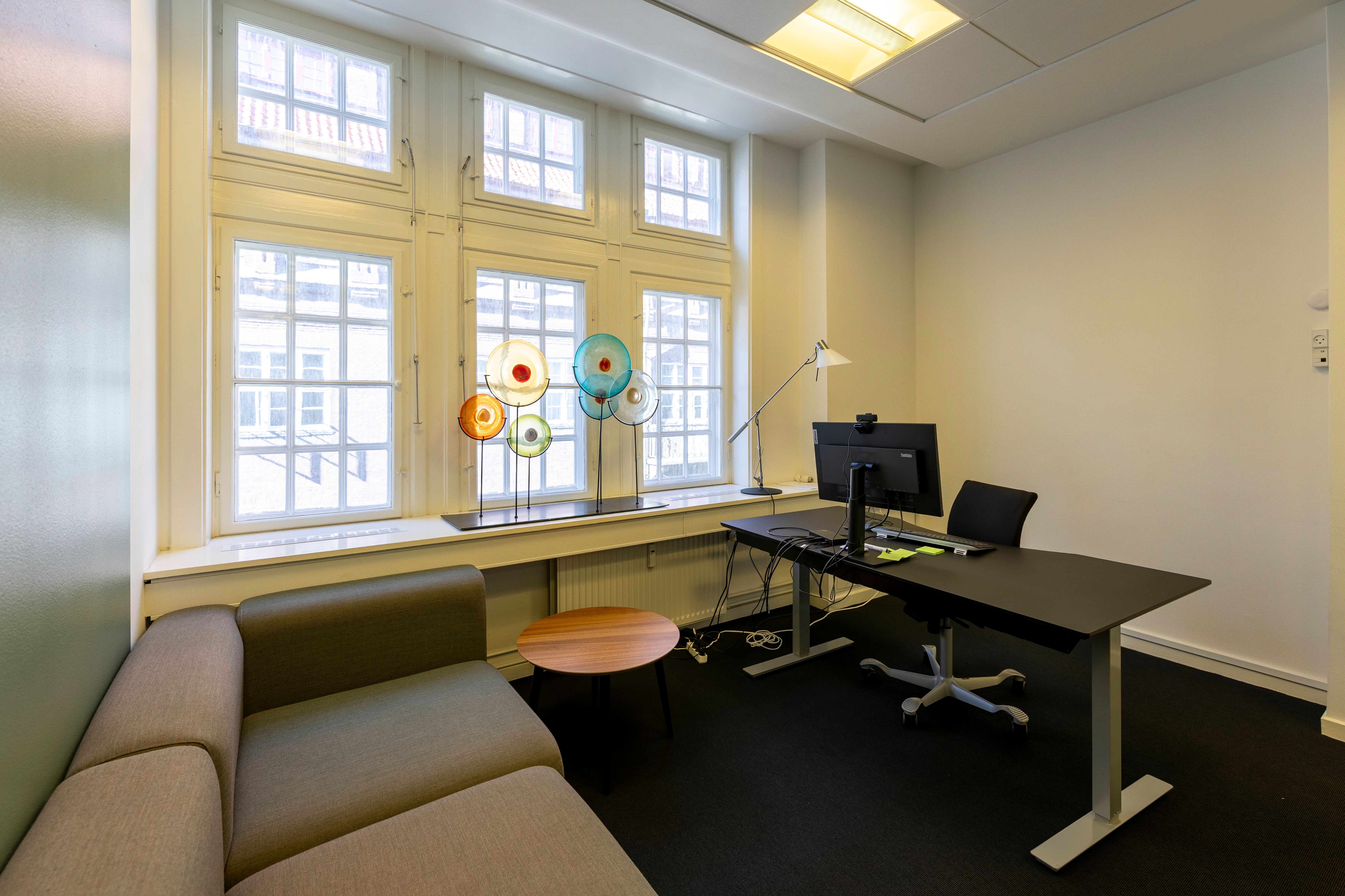 Kontorrum med sofa, skrivebord, stol, skrivebordslampe, computer og farvede glasstykker på vindueskarmen.