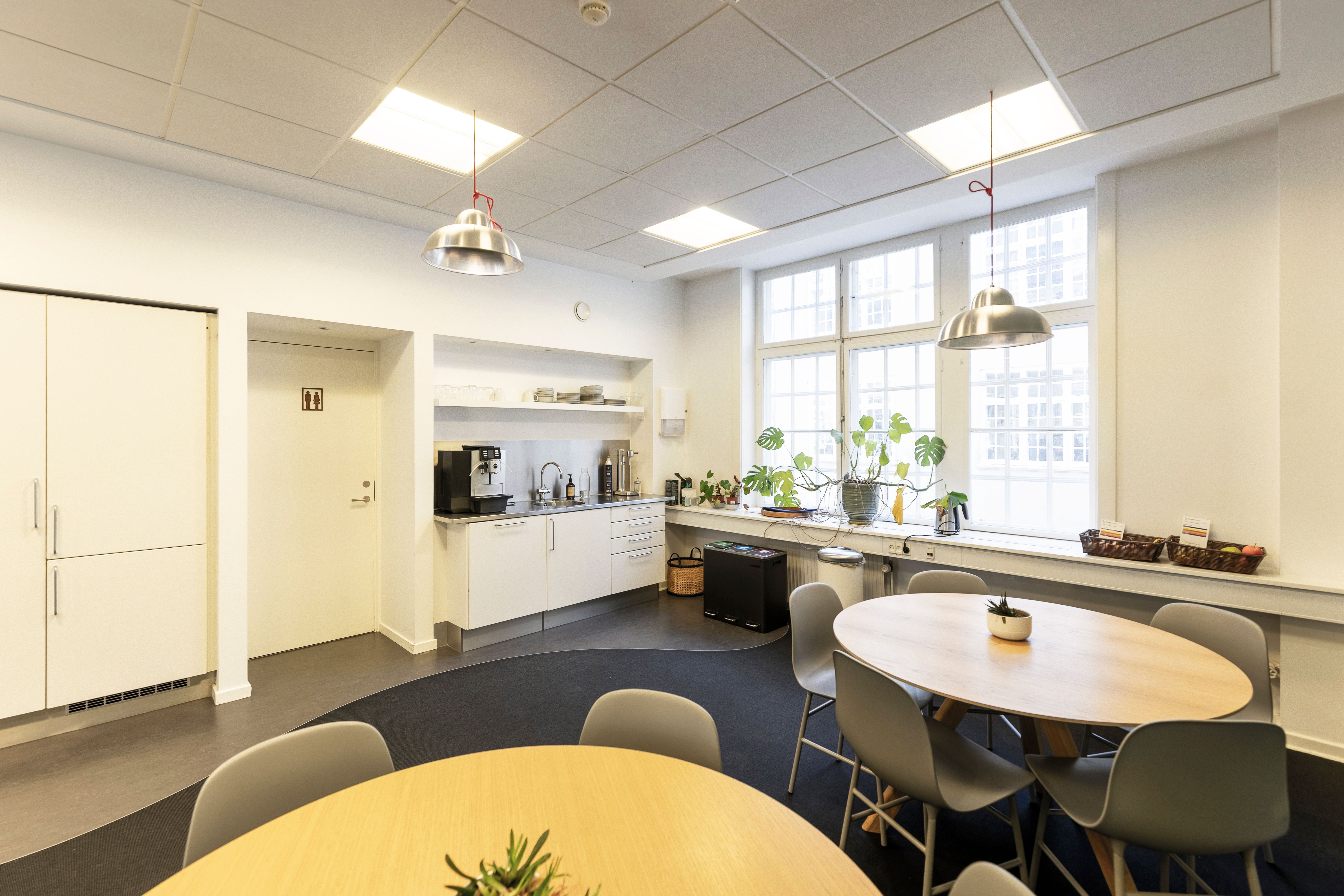 Moderne kantine med lyse loftslampe, hvide vægge, runde træborde, lyse plastikstole, blomster, spisestue med kaffemaskine og krydderier