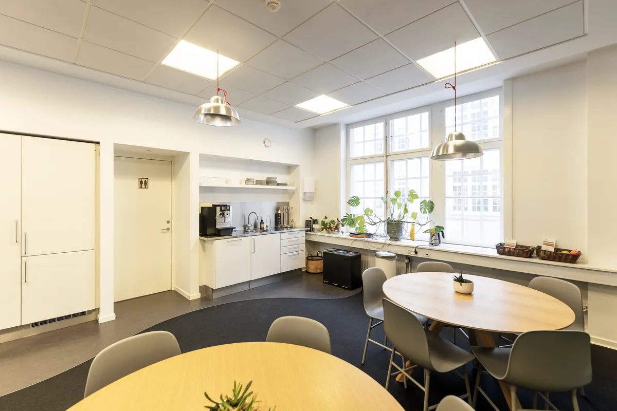 Moderne kantine med lyse loftslampe, hvide vægge, runde træborde, lyse plastikstole, blomster, spisestue med kaffemaskine og krydderier