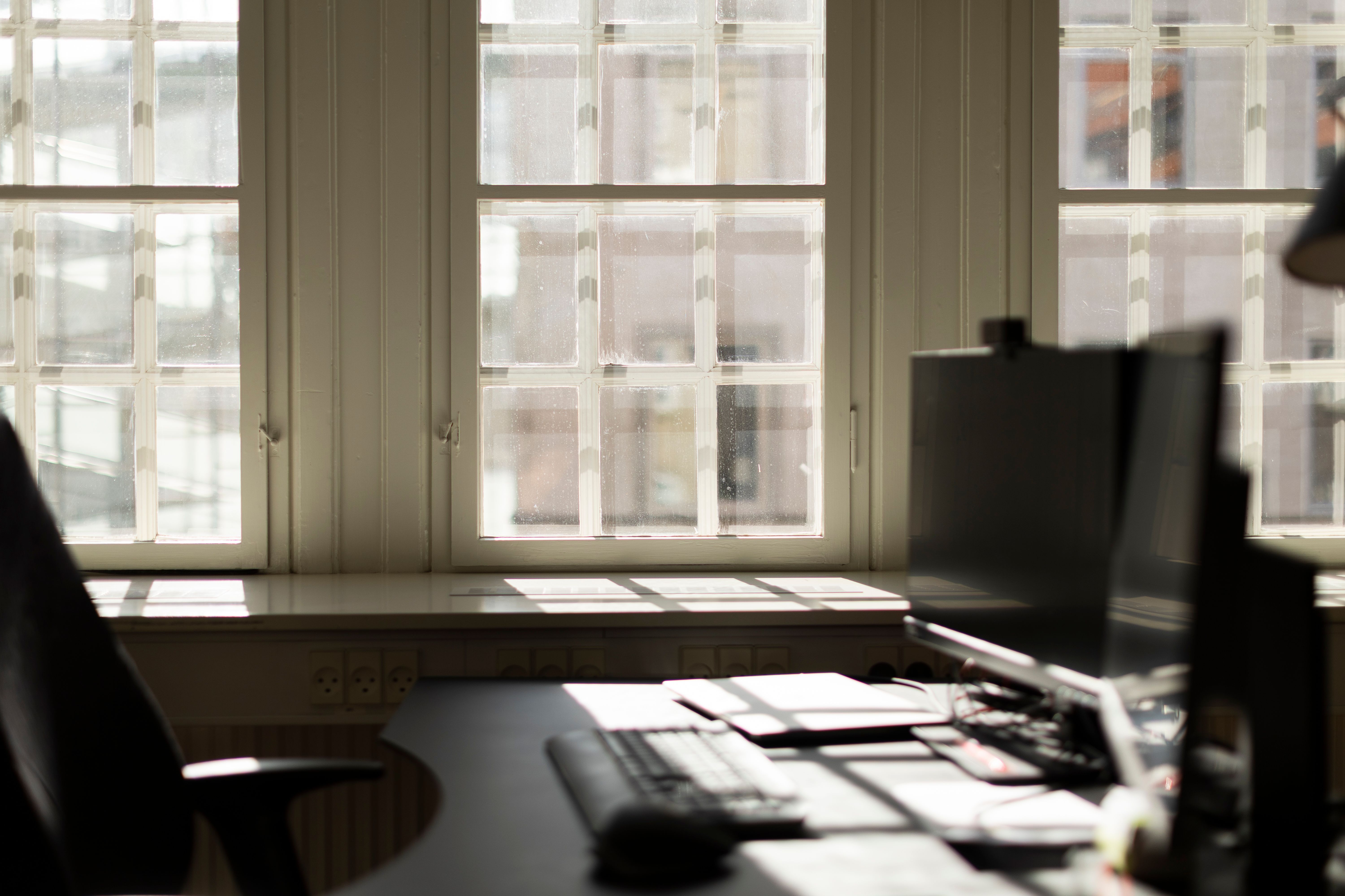 Kontor med to skærme, tastatur, mus, og vinduer med udsigt til Kongens Have. Solskin trænger ind gennem vinduer og lyser op på bordpladen.