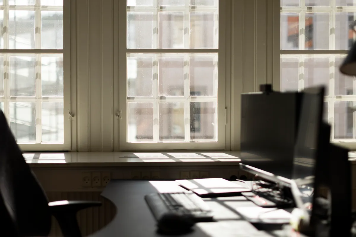 Kontor med to skærme, tastatur, mus, og vinduer med udsigt til Kongens Have. Solskin trænger ind gennem vinduer og lyser op på bordpladen.