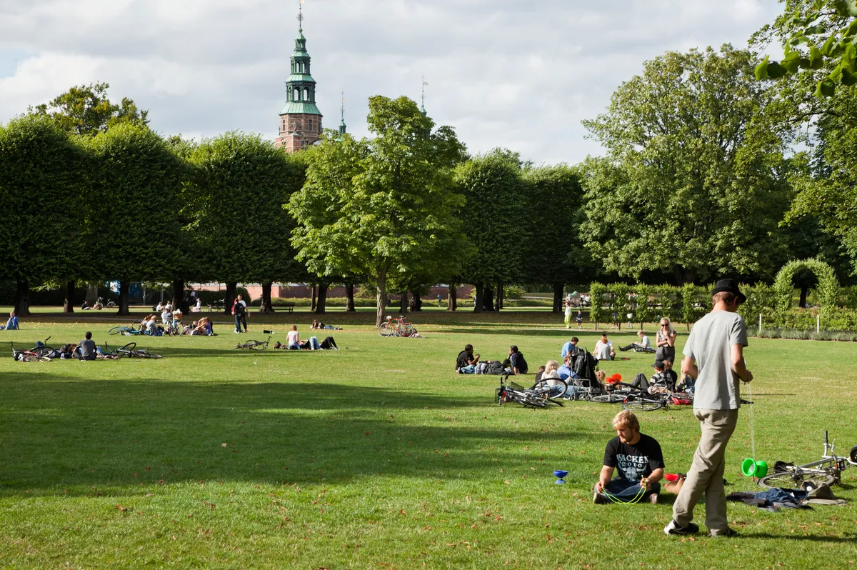 Mange mennesker nyder dagslys på græsfløden i Kongens Have med smukke træer og bygninger i baggrunden
