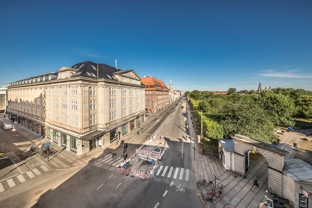 Store bygninger og grønne træer langs en gågade i København. Strømlinede biler og cykler på vejen og mennesker i solskin.