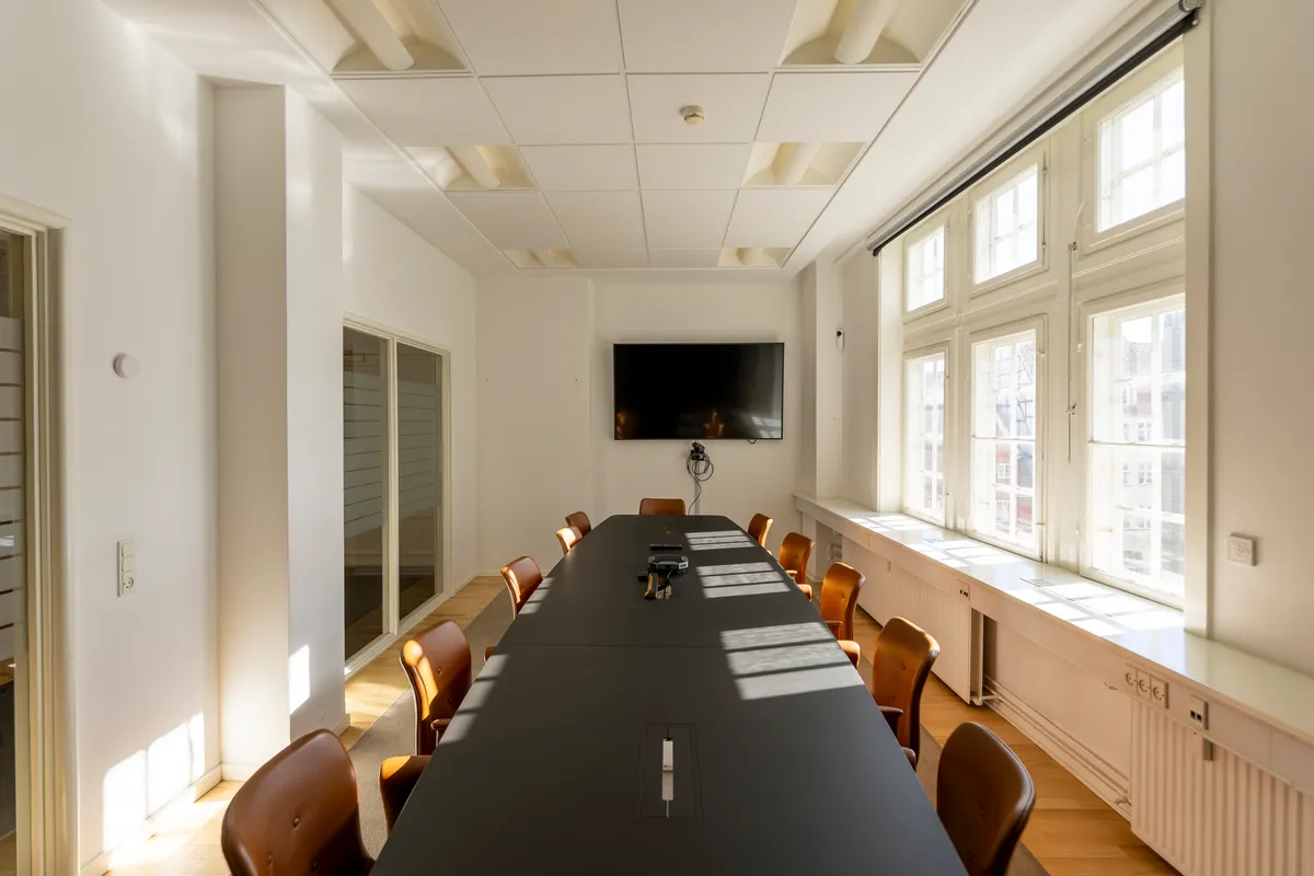 Et klassisk kontorrum med en lang bord og læderstole, en tv-skærm, hvide vægge og trægulve samt store vinduer med solstråler