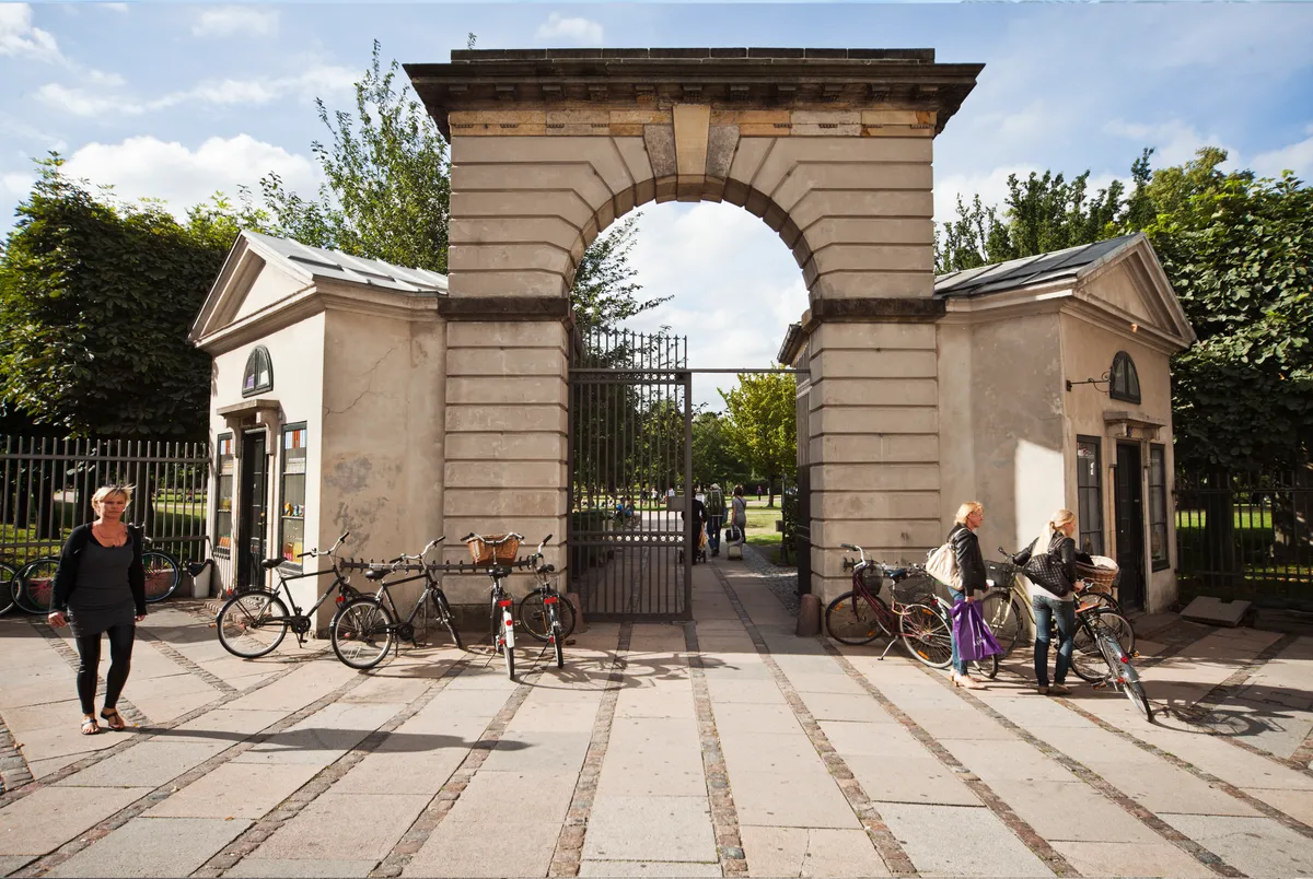 Damer går med deres cykler gennem et stort bueformet indgangsportal, der fører ind i en park. Foran indgangen står flere cykler, og bagved portalen er der et hus og nogle planter.