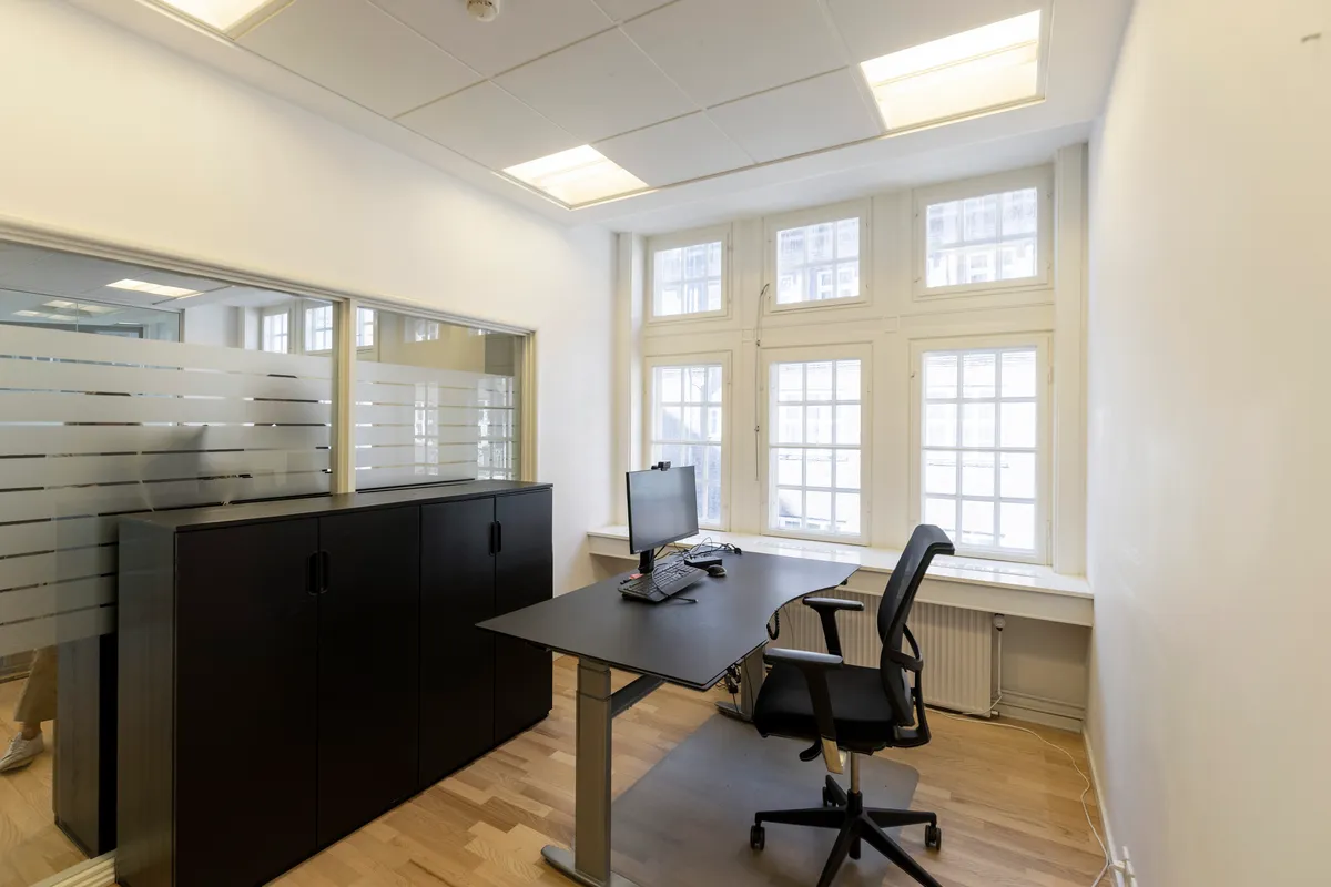 Kontorrum med skrivebord, stol og computer og hvide vægge og loft samt store vinduer med udsigt til byen og Kongens Have