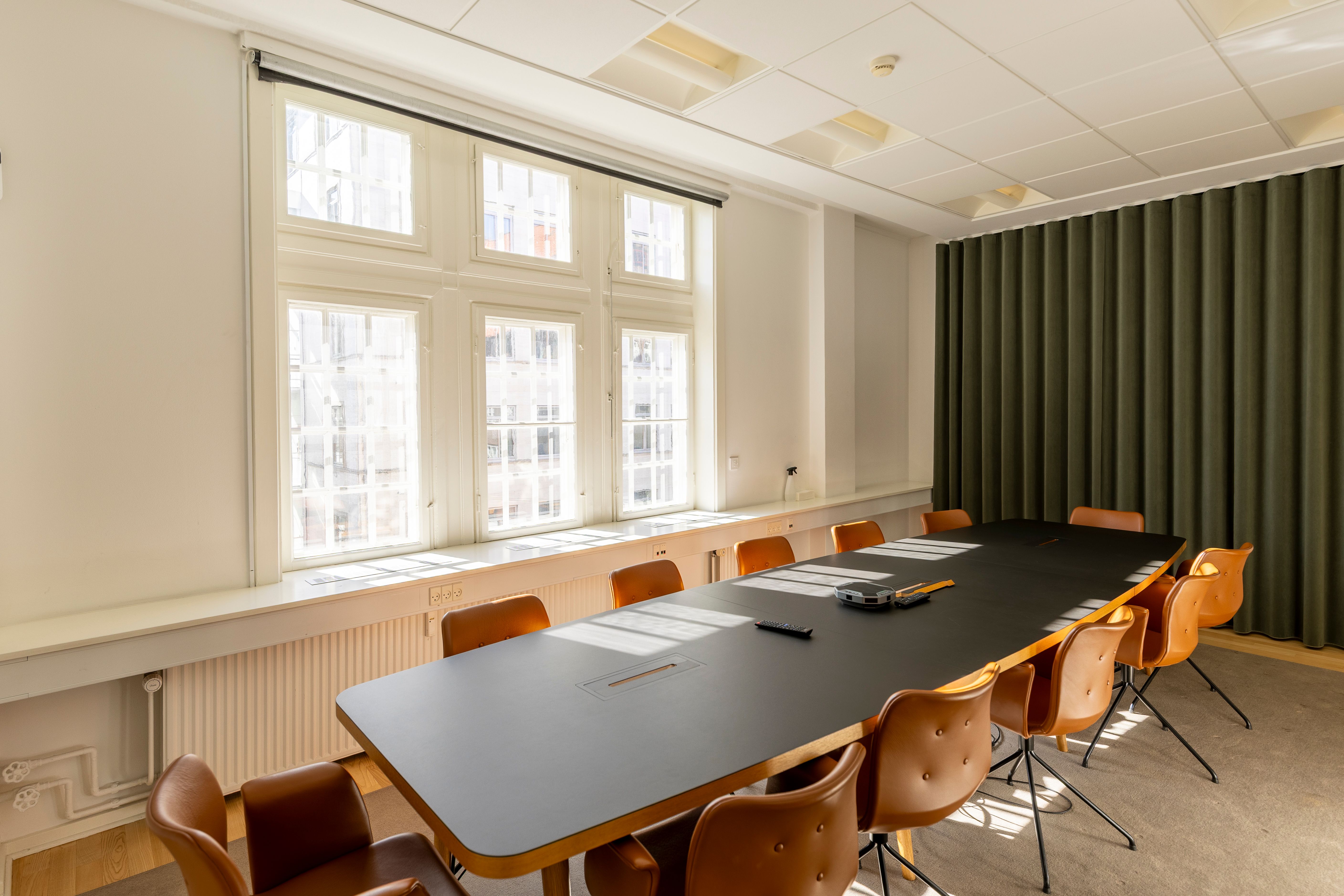 Moderne kontor med lyse vinduer og grønne gardiner, der vender ud mod Kongens Have. Langt, sort bord med brunlæderstole og teknik på bordet.