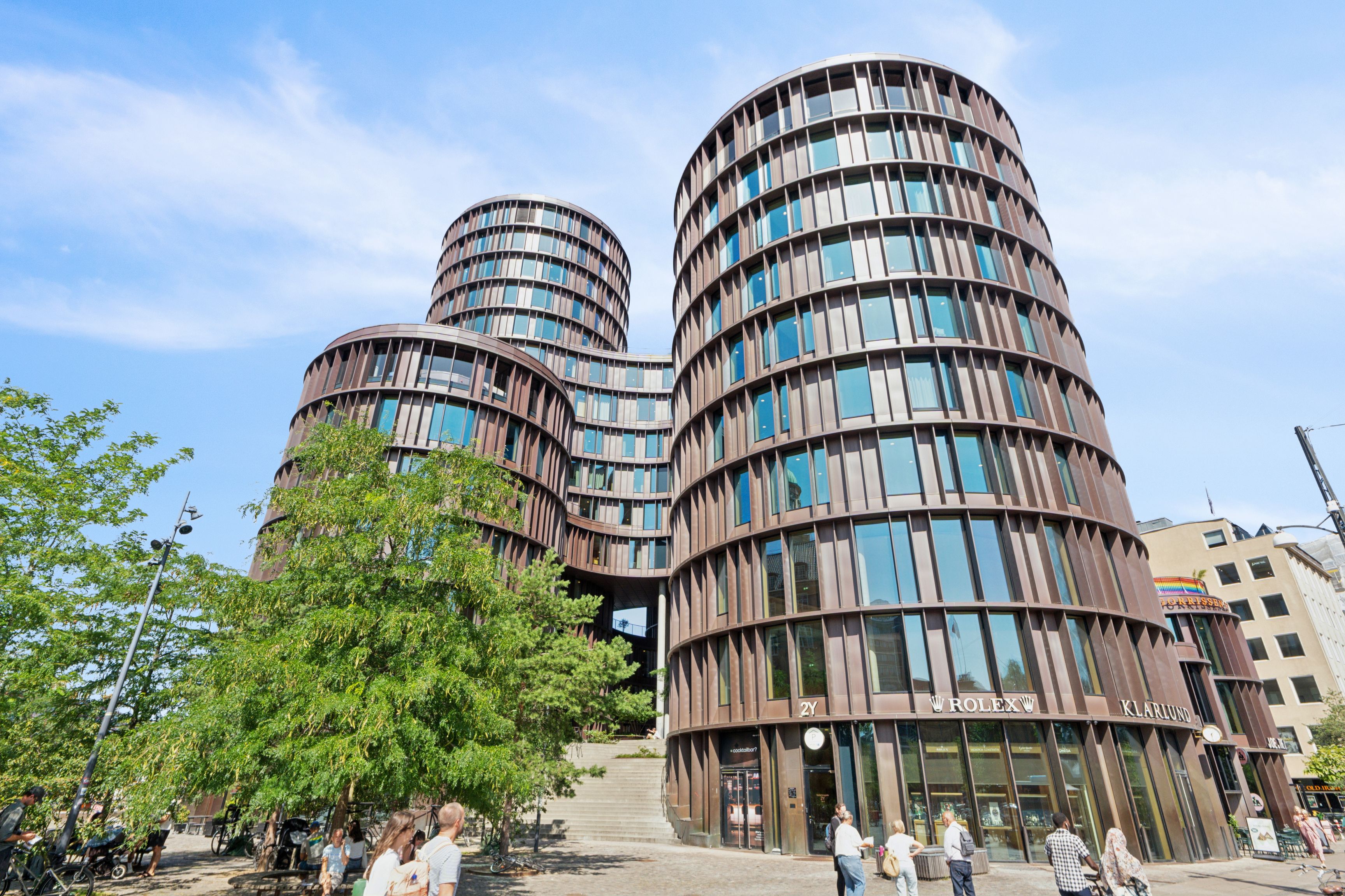 To store, runde bygninger med glasruder og terrasser samt Rolex- og Kjærsgården-skilte. Folk vandrer forbi og parkerer cykler. Store træer og gadevogne.