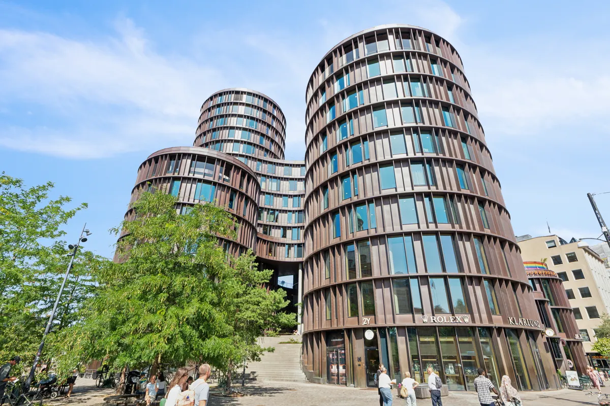To store, runde bygninger med glasruder og terrasser samt Rolex- og Kjærsgården-skilte. Folk vandrer forbi og parkerer cykler. Store træer og gadevogne.