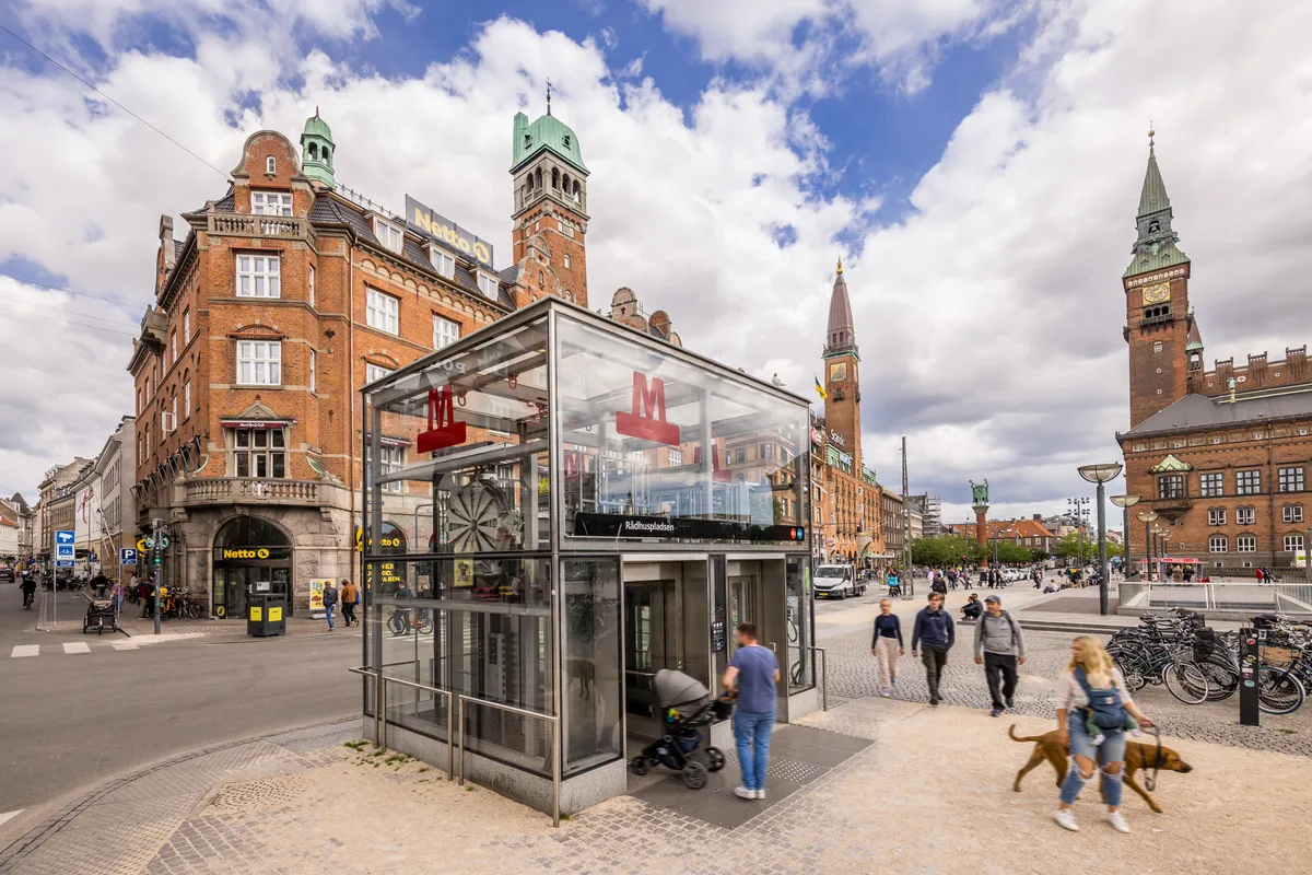 Personer går forbi en busstoppested, der er beskyttet af et glashus med mærket M. Bygningerne i baggrunden har røde mursten og grønne tag. Det er en solrig dag med delvist skyet himmel