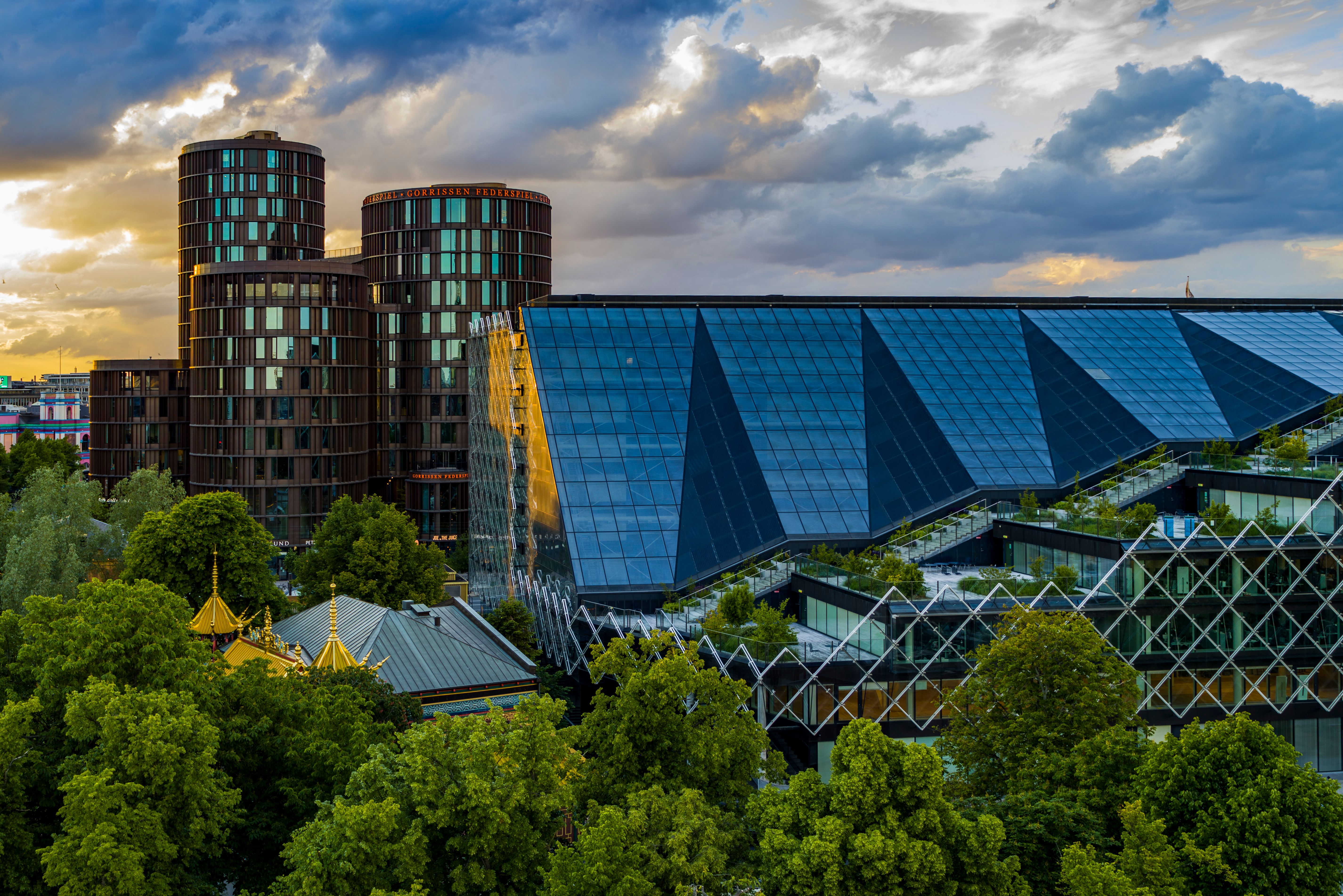 Kontorbyggeri med glasfassade og grønne tag, højde og lyseruder, lys og skyer i baggrunden, smuk udsigt og skov med frodige træer