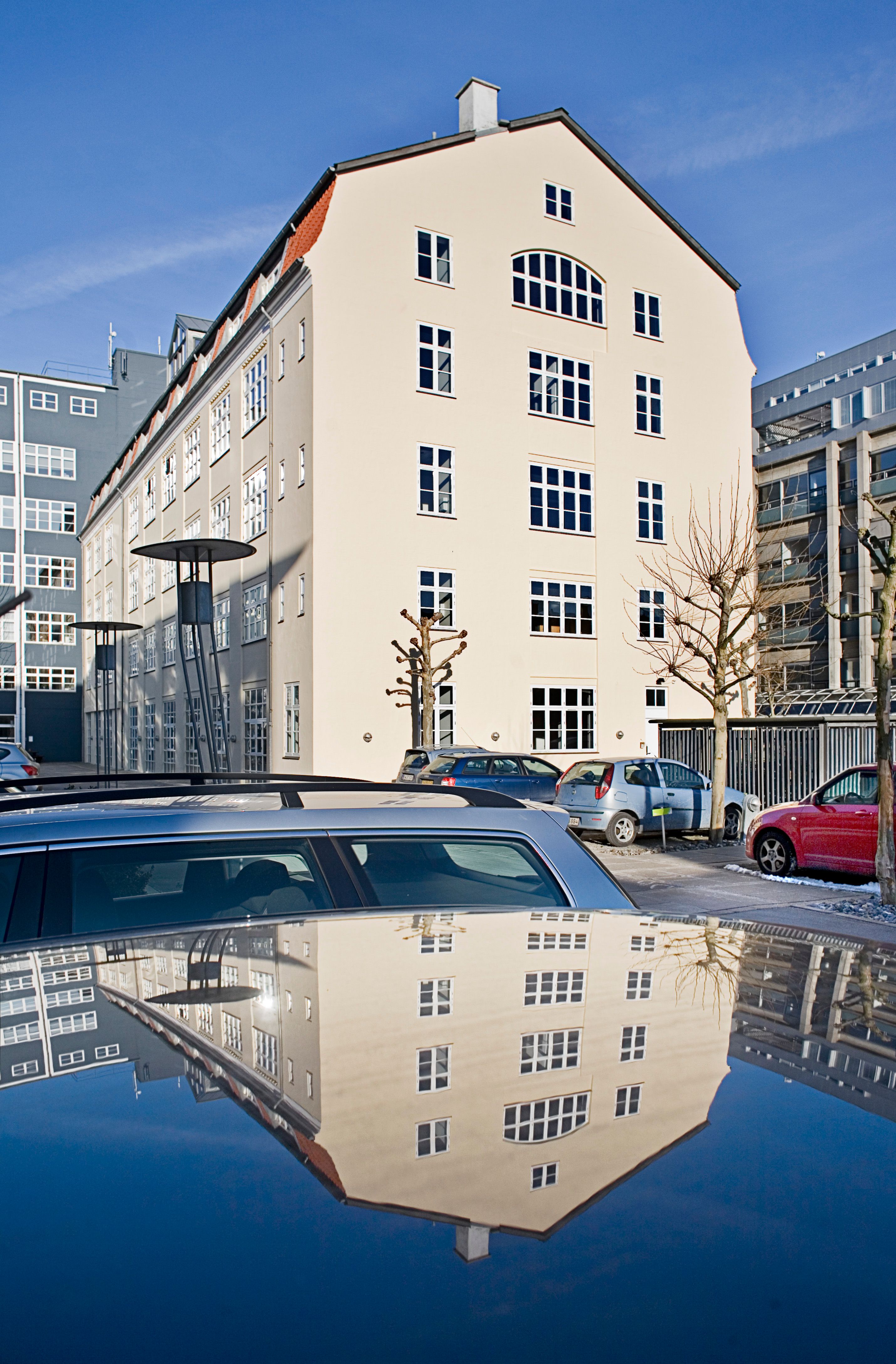 Kontorbygning med lyse vinduer og røde tagkanter. Biler parkeret på en parkeringsplads. Træer og en lysstoftlampe på gaden. Refleksion af bygningen i den plettede vandoverflade.