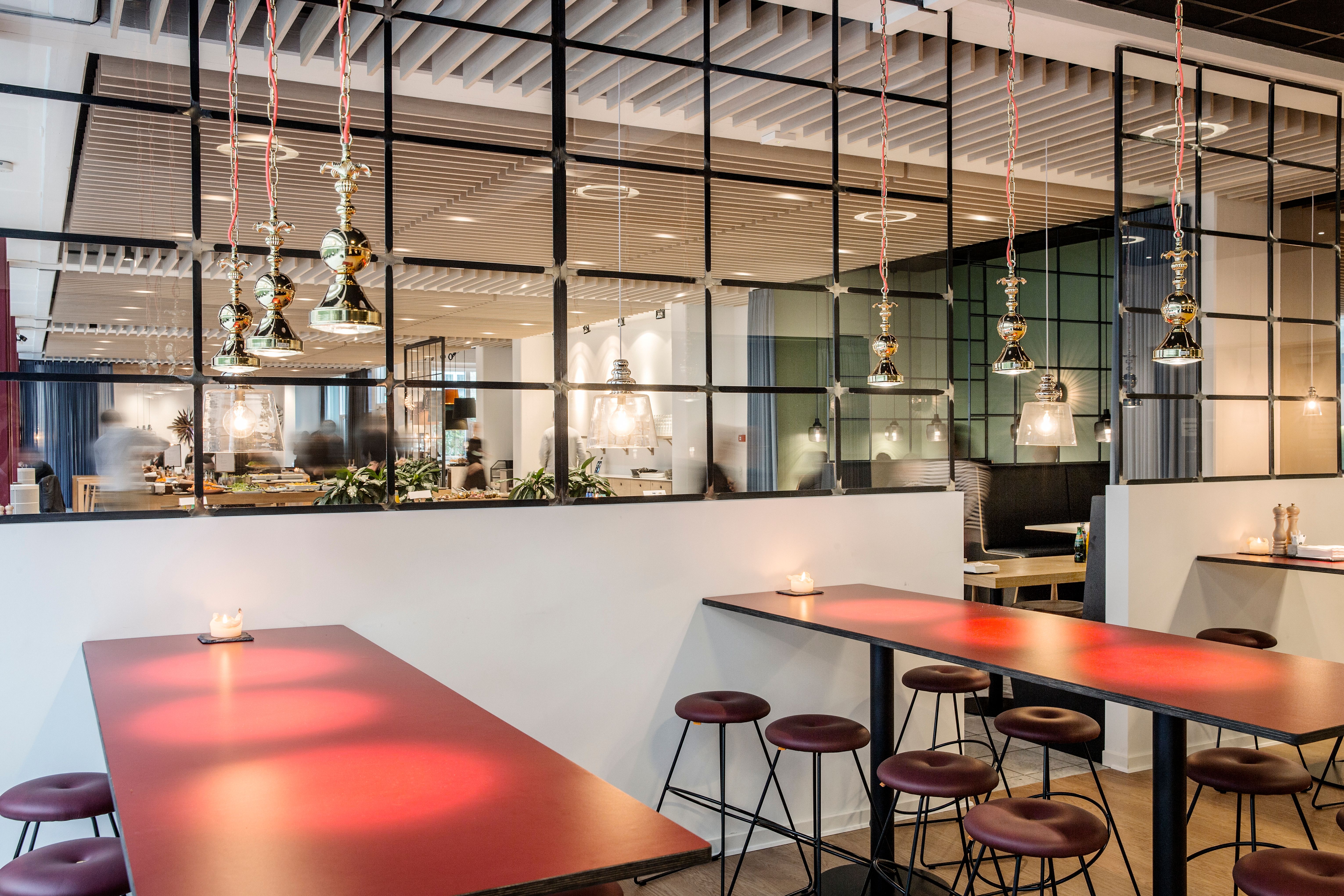 Indenfor en elegant, højloftet lokale med røde borde og stolpe, hænger lysekroner og reflekterer lys fra stearinlys. Store spejle reflekterer rummet, og der er grønne planter i baggrunden.