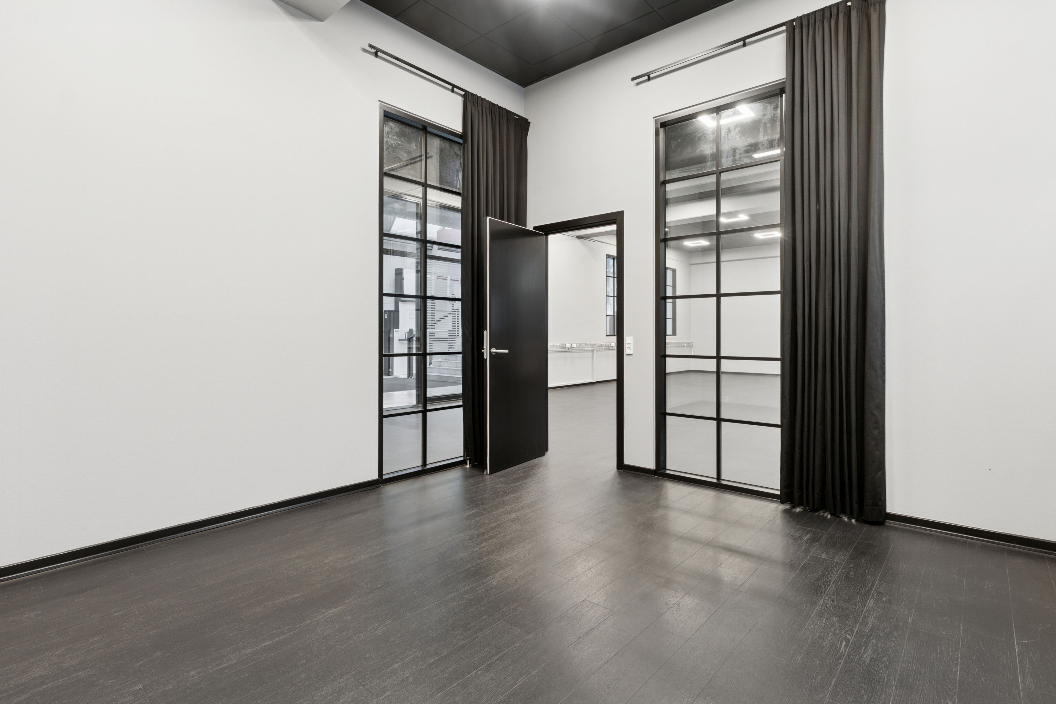 Minimalistisk indretning med højloftede rum, to åbne glasdøre og store vinduer, der giver masser af lys. Sorte vægge og gulv samt store sortbrune gardiner.