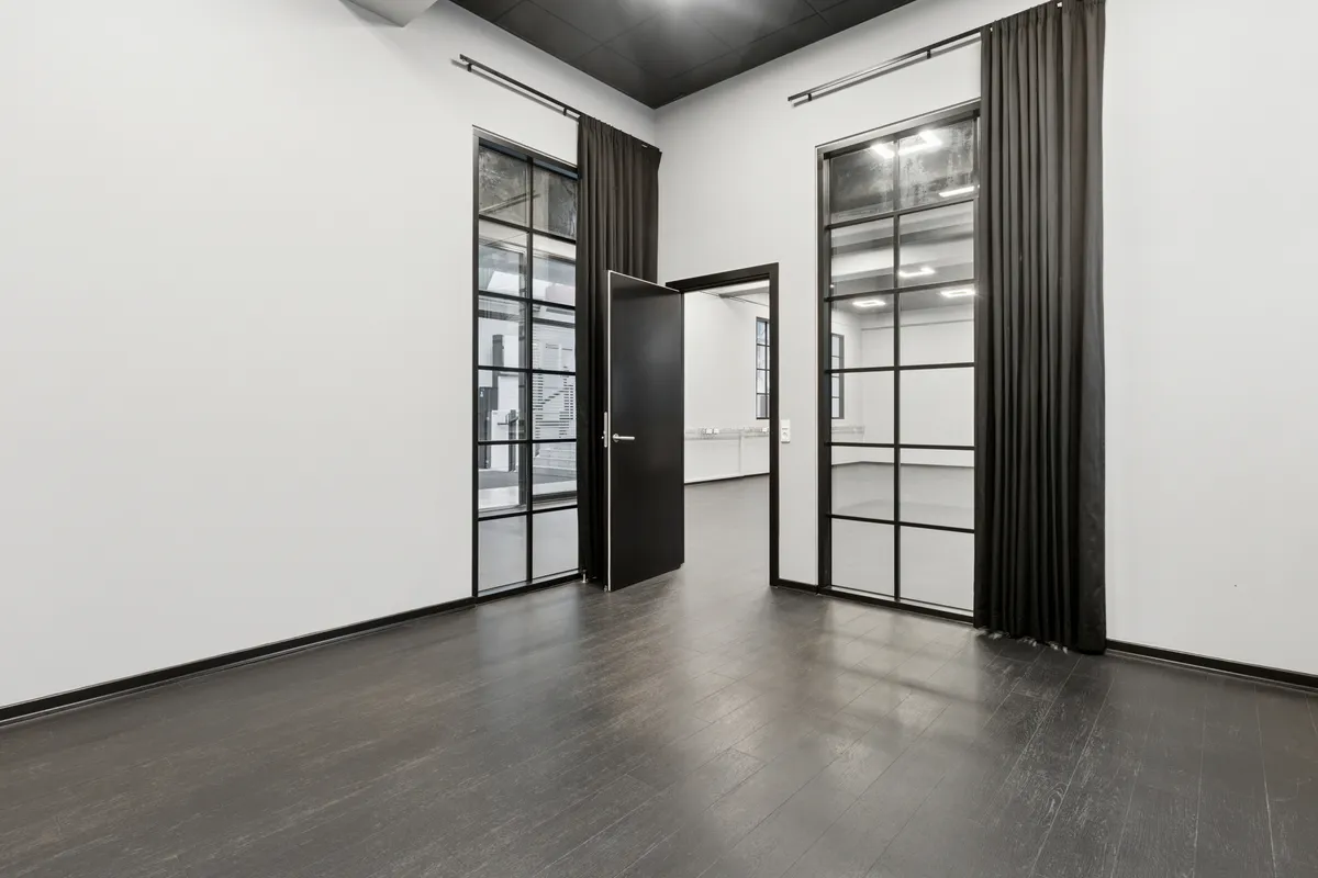 Minimalistisk indretning med højloftede rum, to åbne glasdøre og store vinduer, der giver masser af lys. Sorte vægge og gulv samt store sortbrune gardiner.