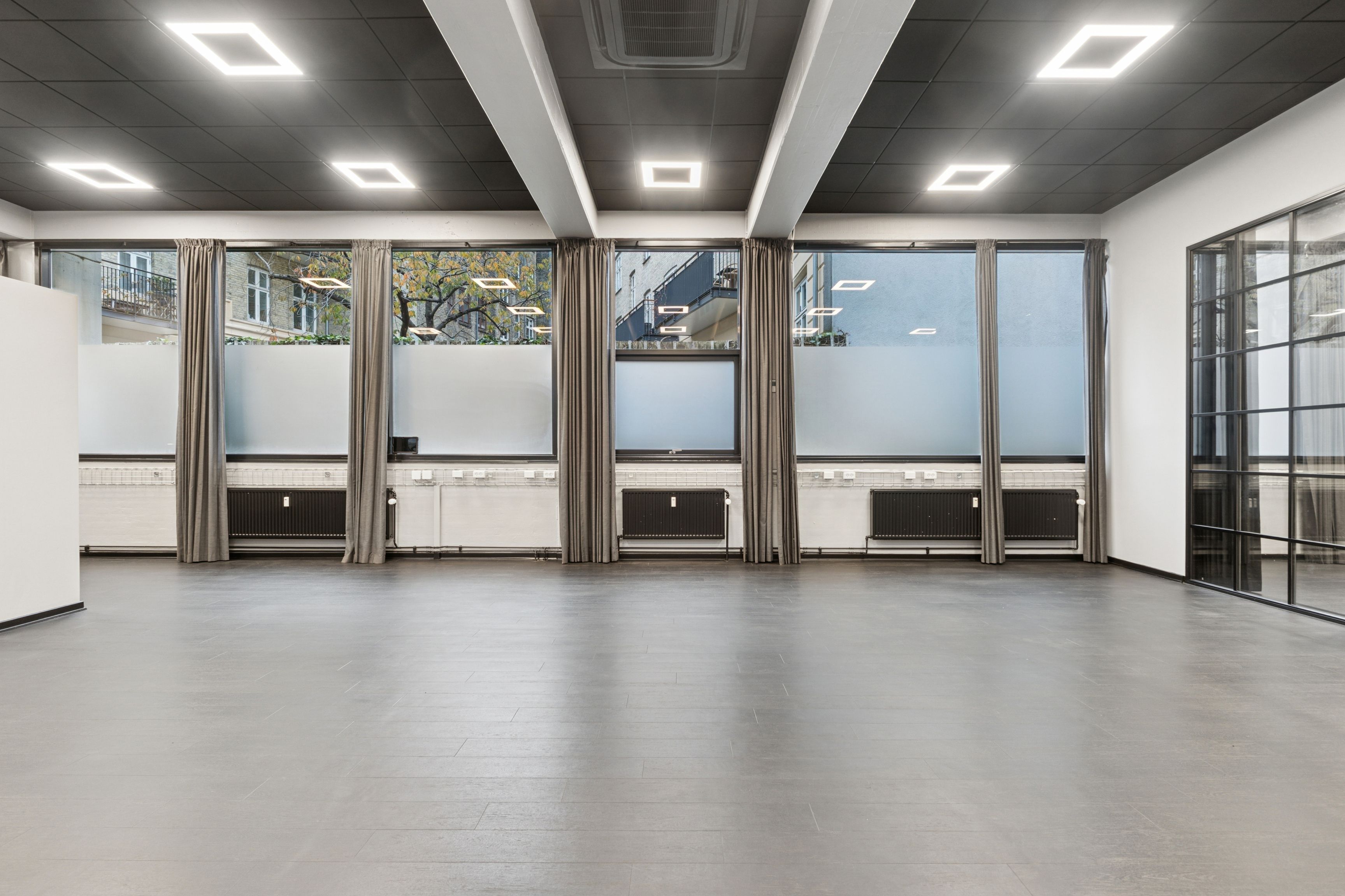 Lejlighed med stort rum, lyse loftsvinduer og slanke hjørner. Væggen er hvid og gulvbelægningen er grå. Vinduerne er store og udstyret med skyggegardiner