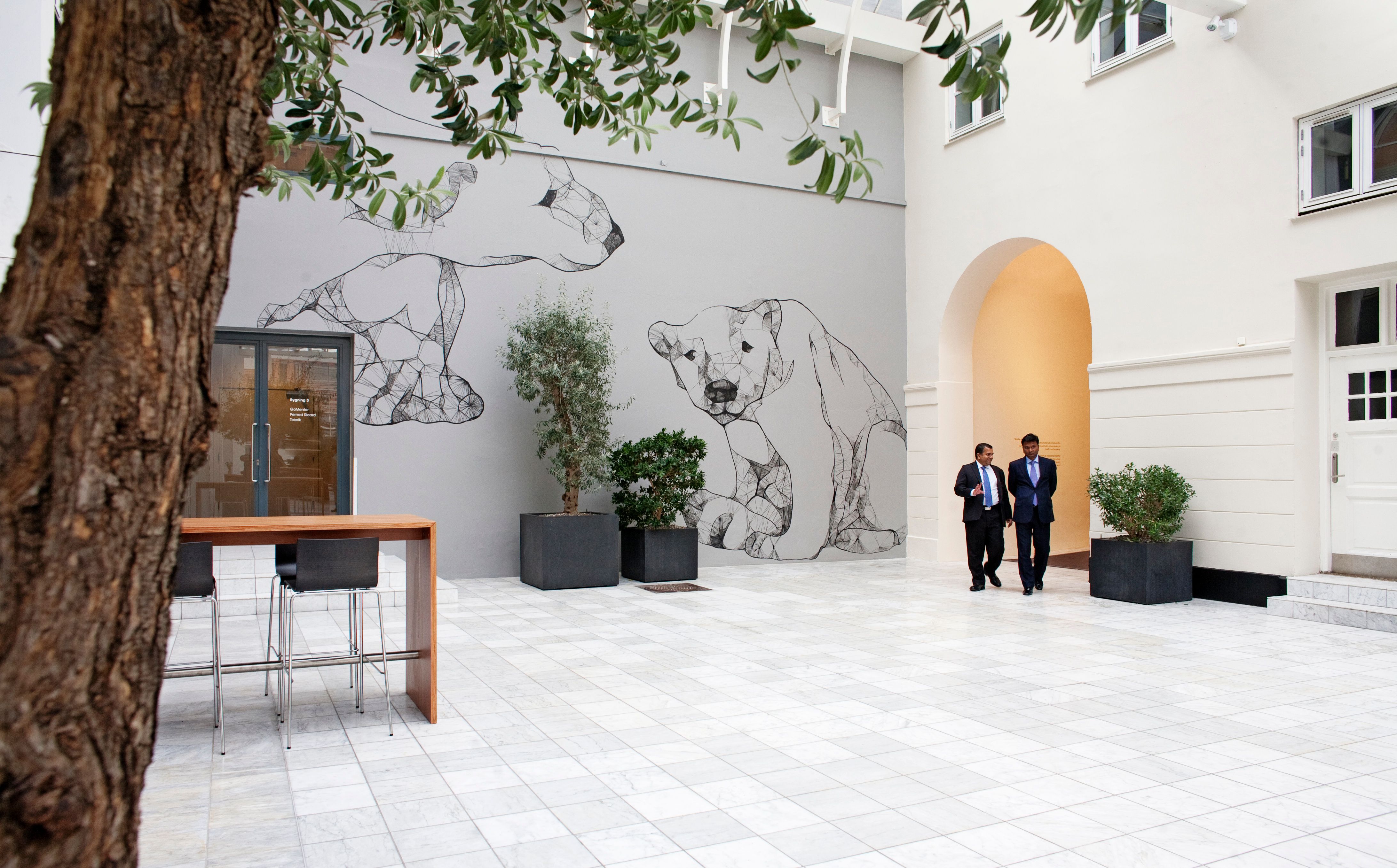 To forretningsfolk går gennem en gårdhave med marmorgulv, grafisk vægmaleri af bjørne og lyse vinduer på højloftet bygning