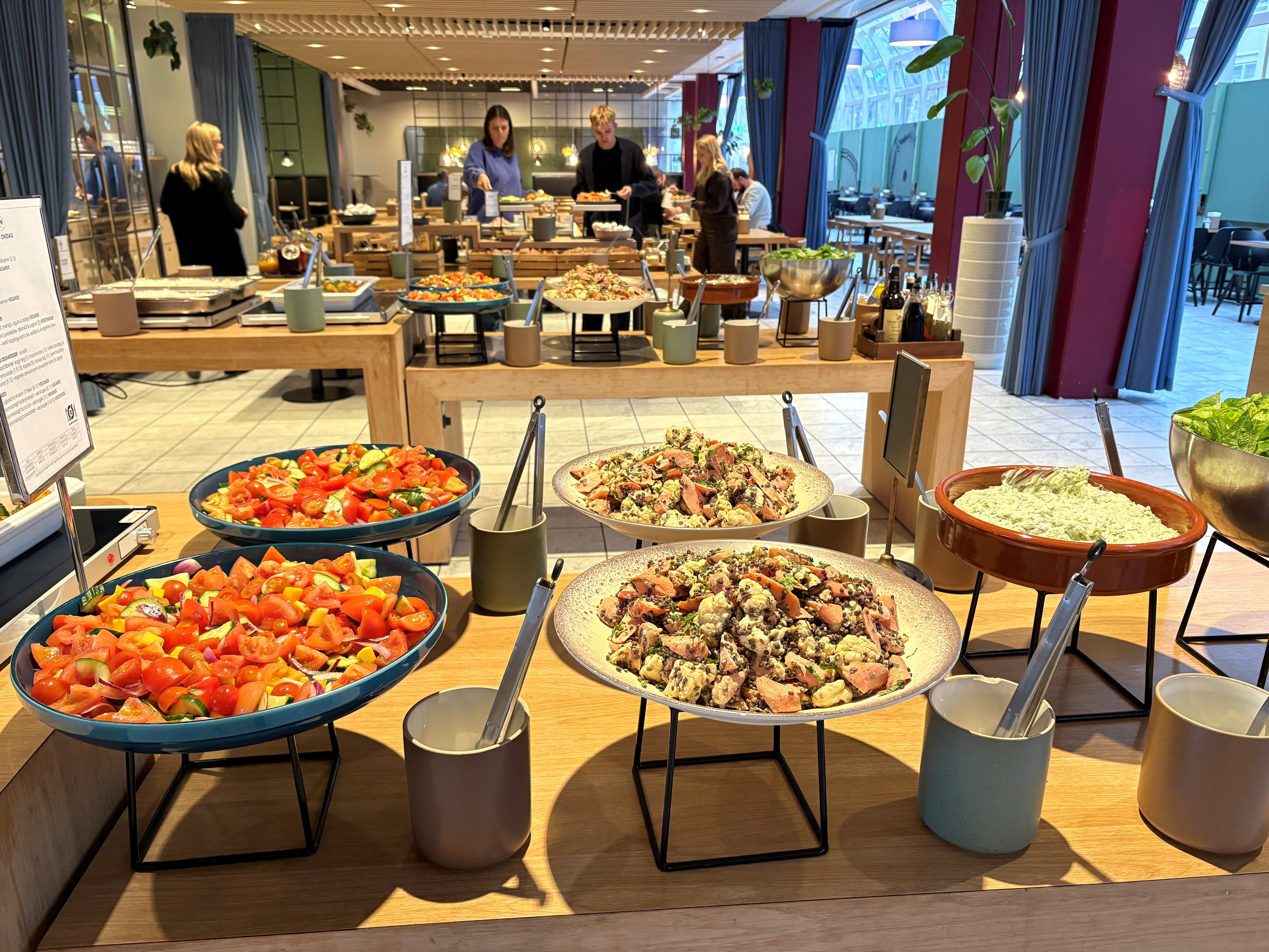 Buffet med diverse retter og salater samt serveringsudstyr og røde vægge. Buffeten er indrettet med store, runde tallerkener og høje serveringsstativer.
