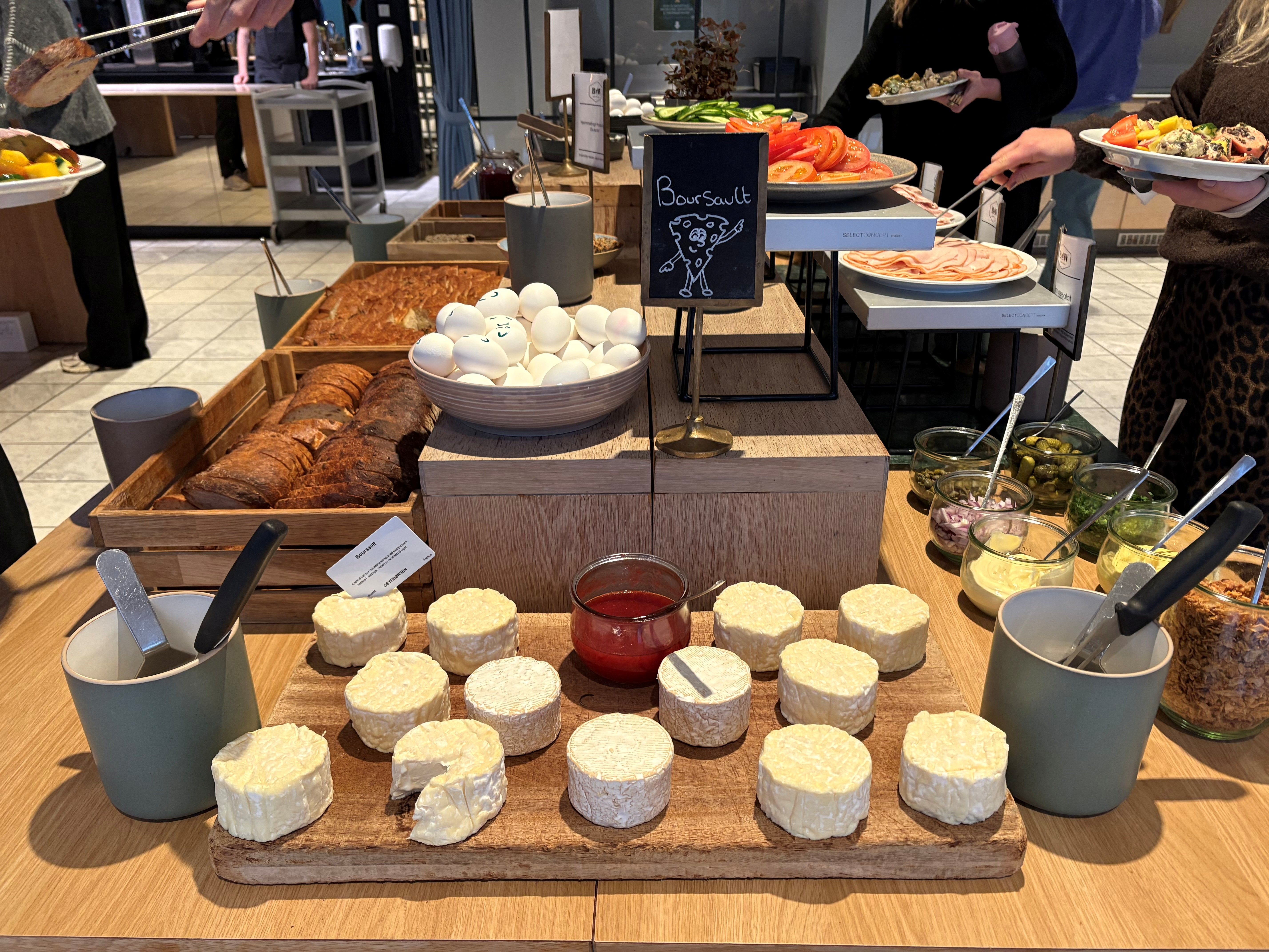 Buffet med frokost, hvor der er serveret røde tomater, grønne salatgrøntsager, hvide æg, friskbrød og runde, hvide ostestykker med rødt sylt og krydderurter