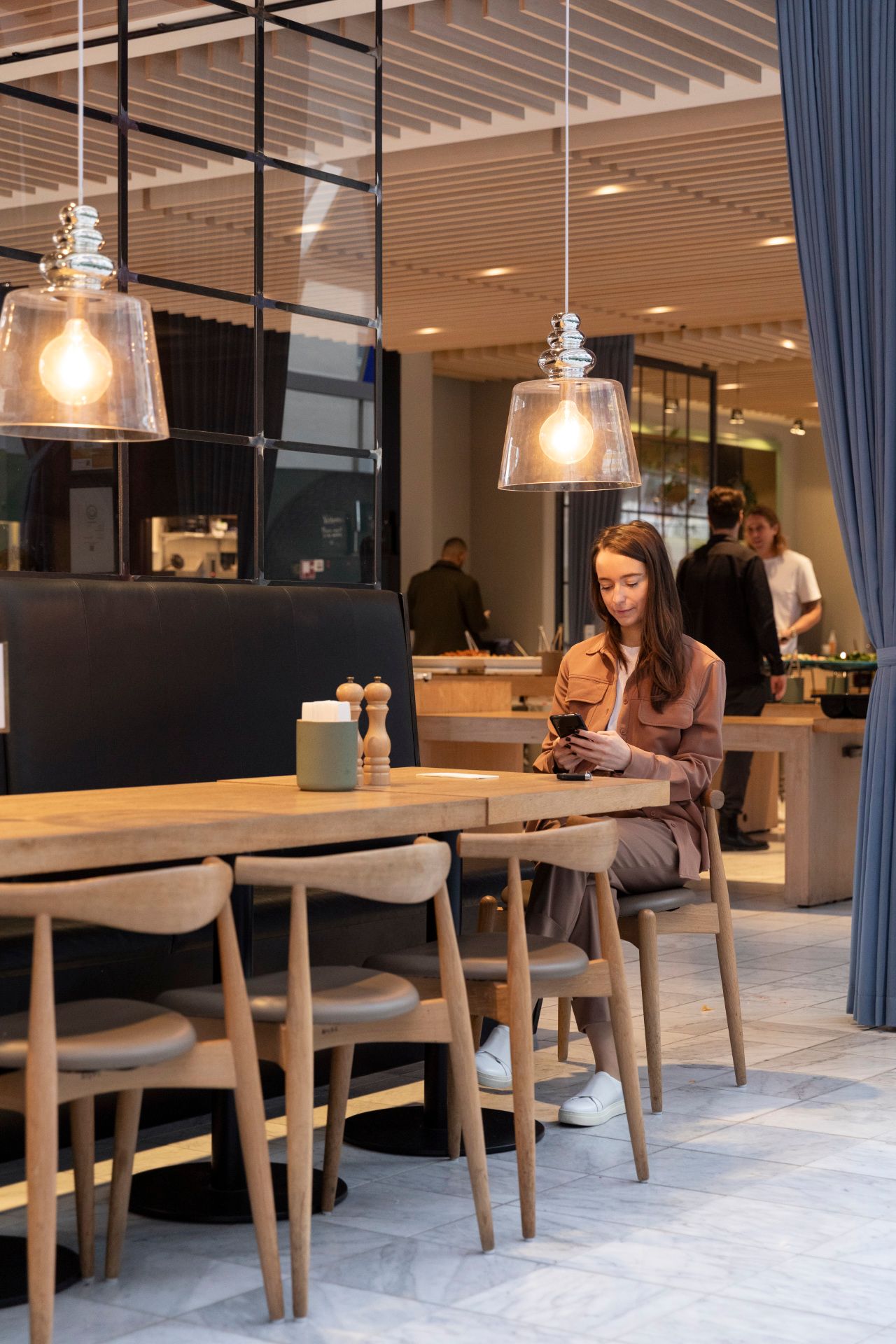 Dame sidder og bruger mobiltelefon ved et rundt bord med tre stole i en møbleret kontorrestaurant med loftsvinduer og lysekroner