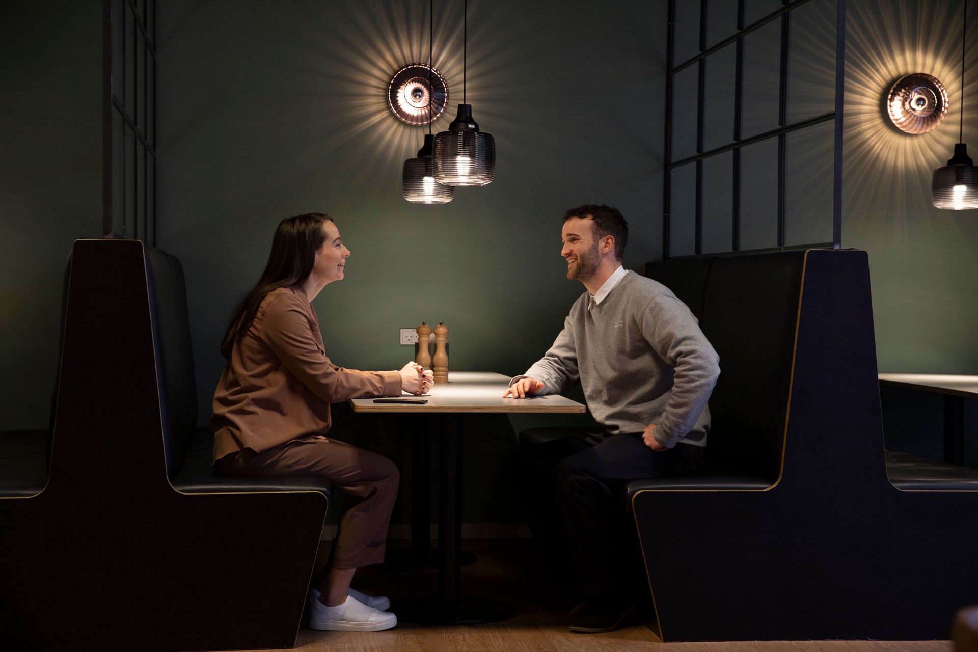 To mennesker i en mørk kontorlounge med højloftede rum og lysekroner med spejle i loftet, der reflekterer lys på væggen