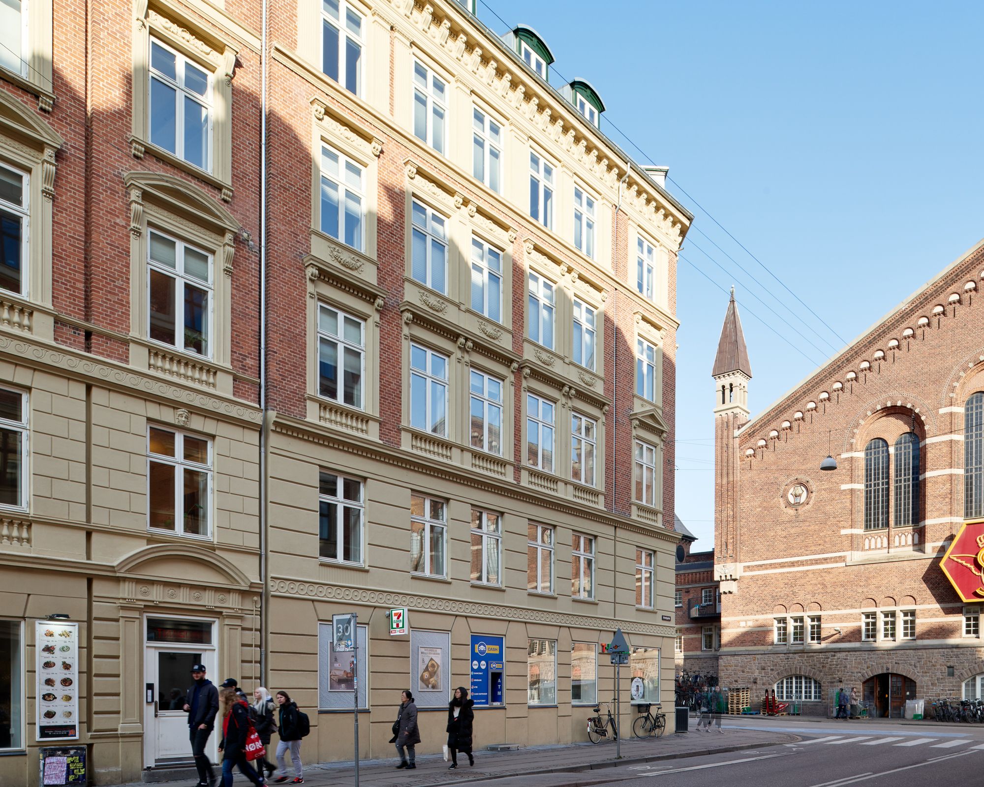 På Reventlowsgade 14, i København, står et nyrenoveret kontorbygning med røde mursten og store vinduer. Folk går forbi og en 7-Eleven ligger på gadehjørnet