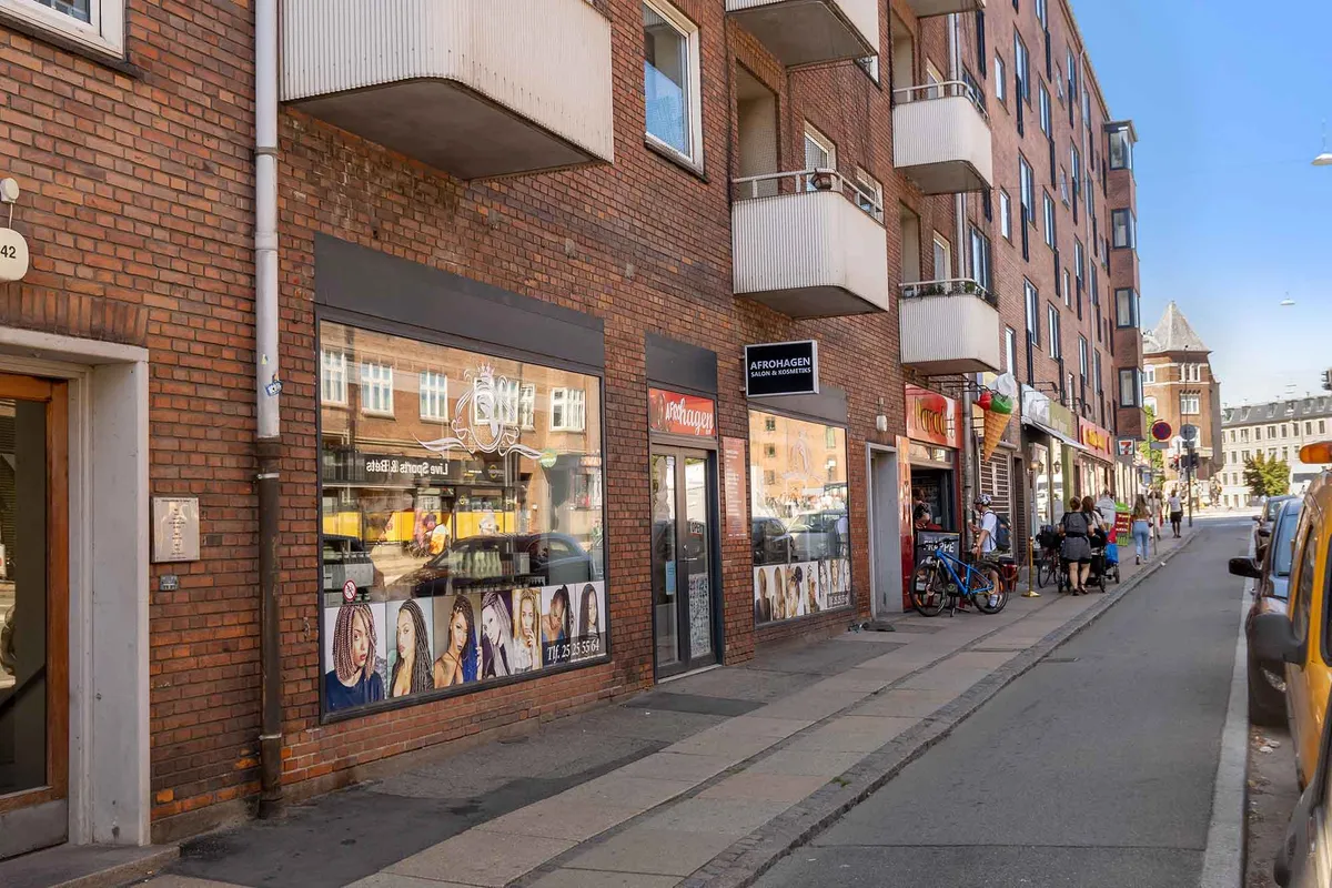 Butikslejemål på Toftegårds Allé med moderne facade og glasvinduer, hvor folk går forbi og nyder slik og drikkevarer i en solrig dag