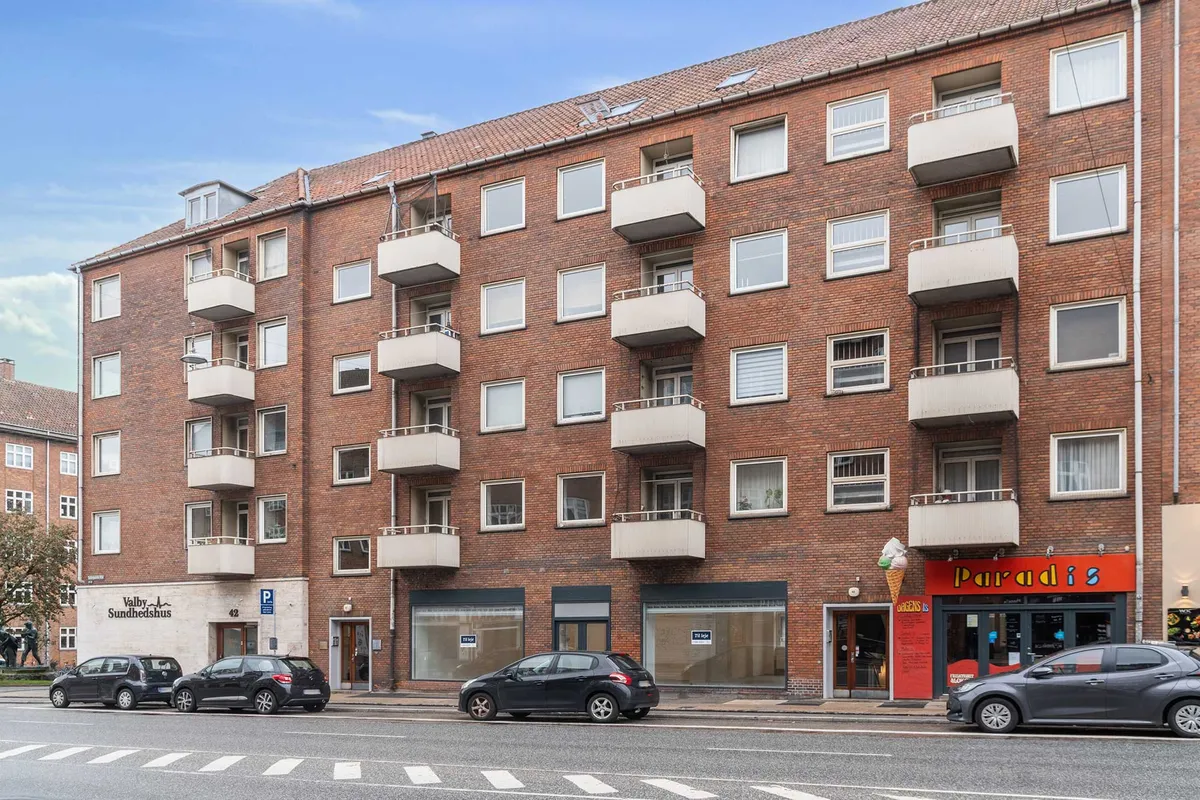 Fire biler parkeret foran et lejemål i Valby. To biler parkeret på hver side af gågaden og to butikker med Paradis is og Valby Sundhedshus på hjørnet