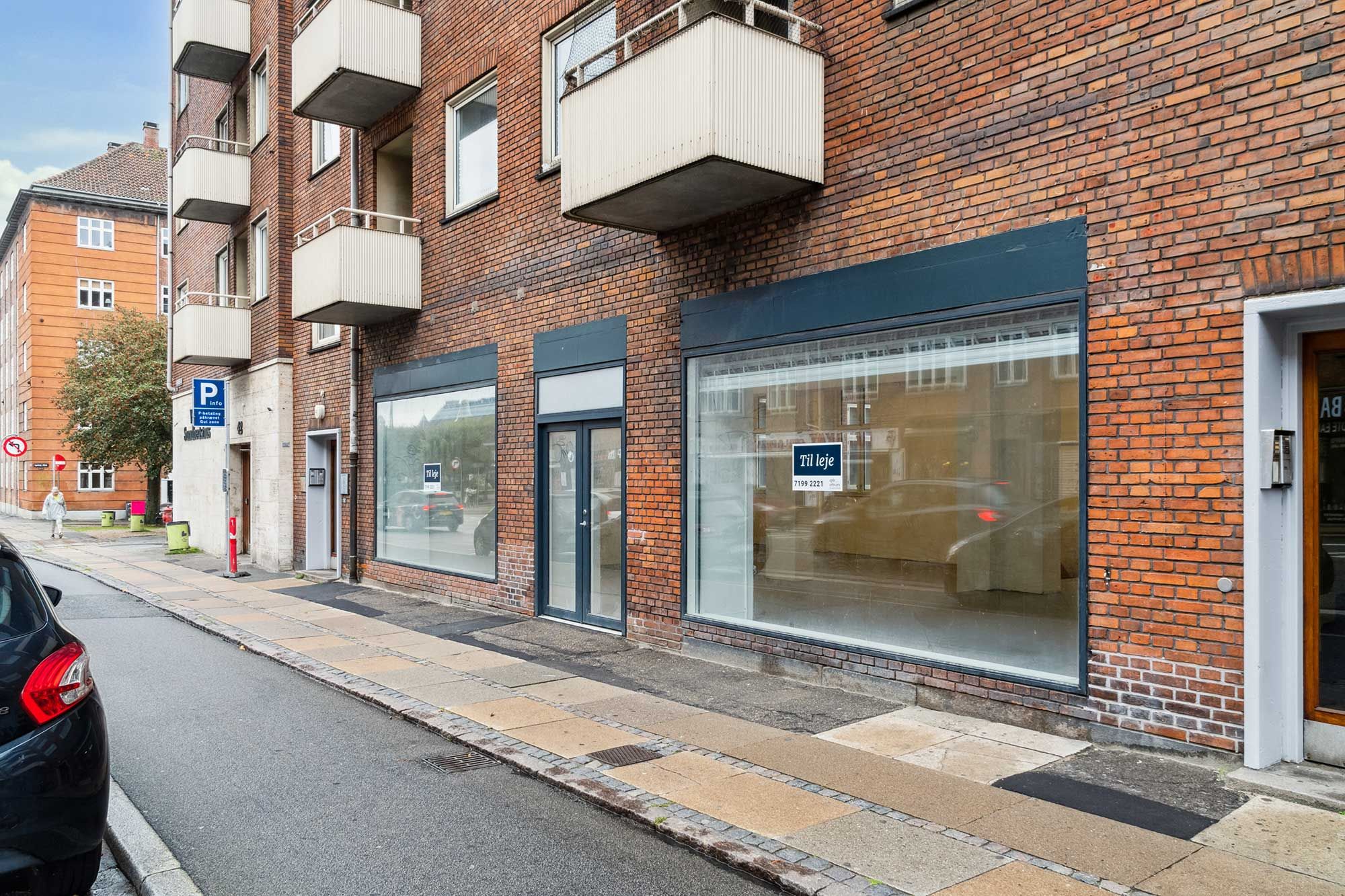 Butikstil leje i Valby med glasvinduer og røde mursten. Til leje-skilt og rødt stoptegn. Parkeret bil og gul belægning på gaden.