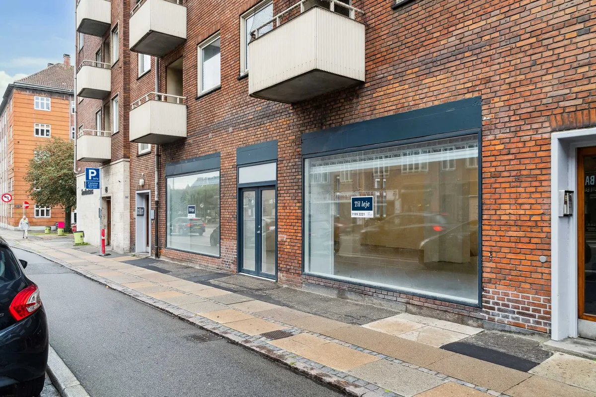 Butikstil leje i Valby med glasvinduer og røde mursten. Til leje-skilt og rødt stoptegn. Parkeret bil og gul belægning på gaden.