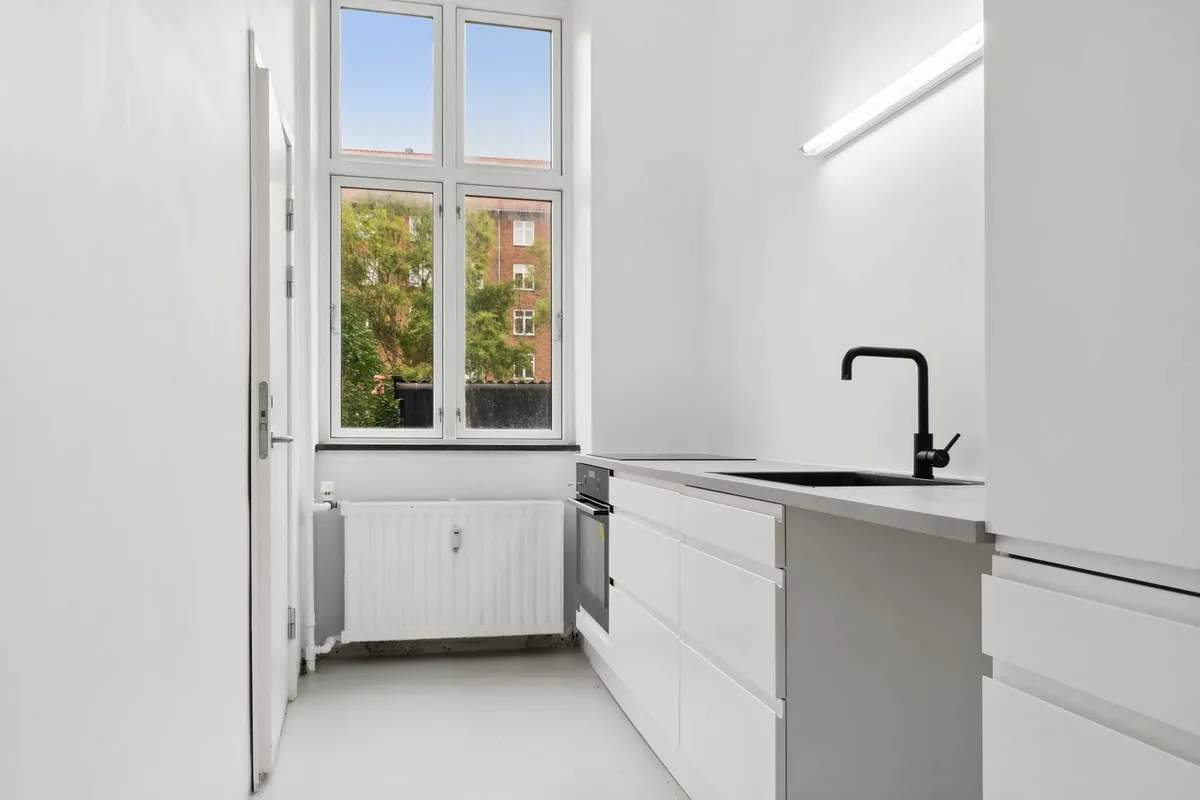 Moderne køkken med hvidt skab, lysekroner og køkkenvæg. Sort vandhane og ovn samt radiator under vinduet. Store ruder med udsigt til grønne træer og bygninger