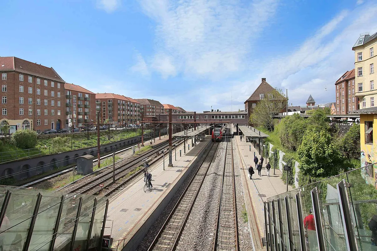 Mennesker går på en togstation, hvor der er to spor med sporvognsskinner og sporvogne. Stationsbygning og grønne planter og træer er i baggrunden.