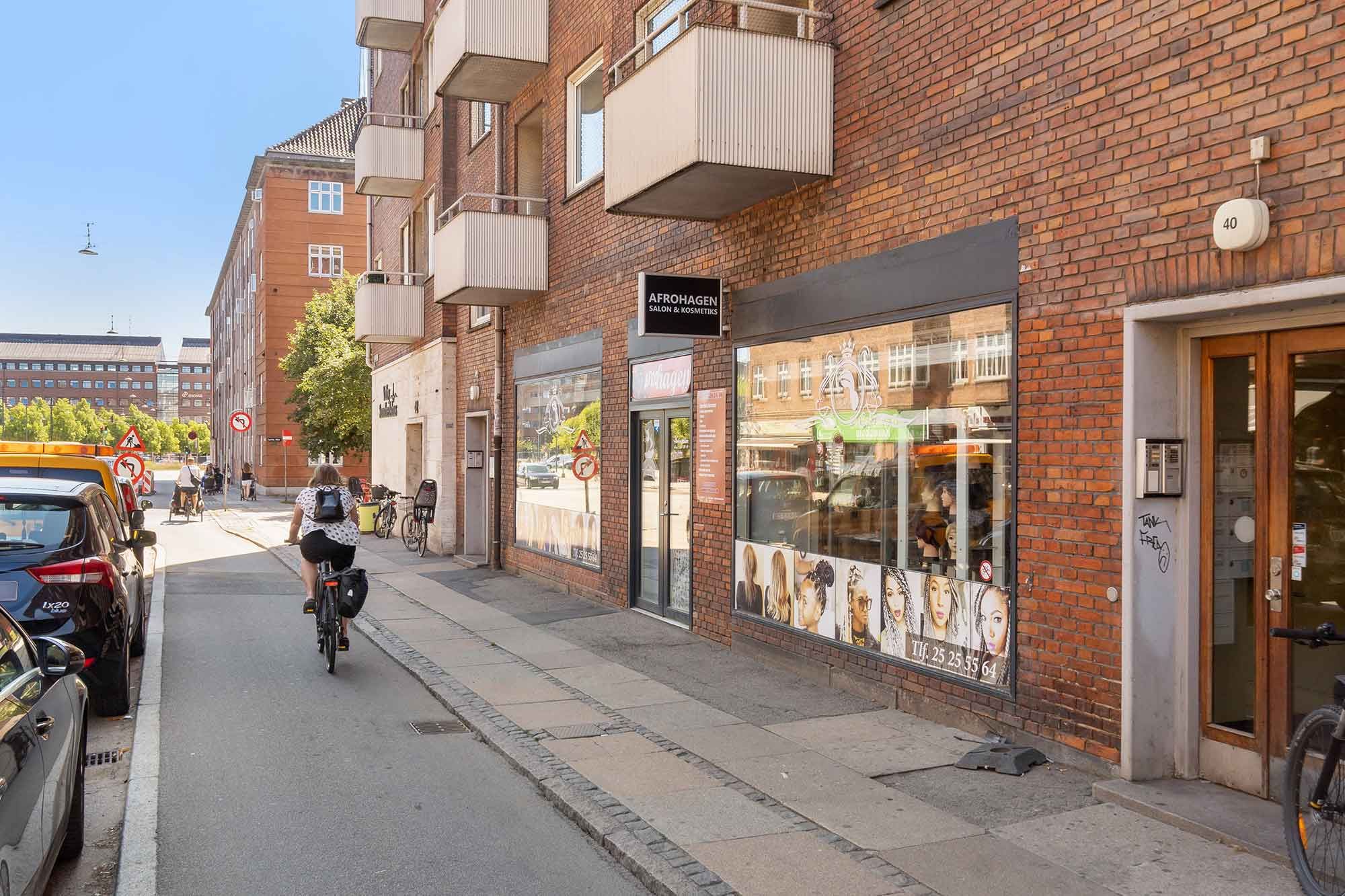 En kvinde kører på en cykel ad en smal gågade i Valby, med en salon på højre side og parkerede biler på venstre side