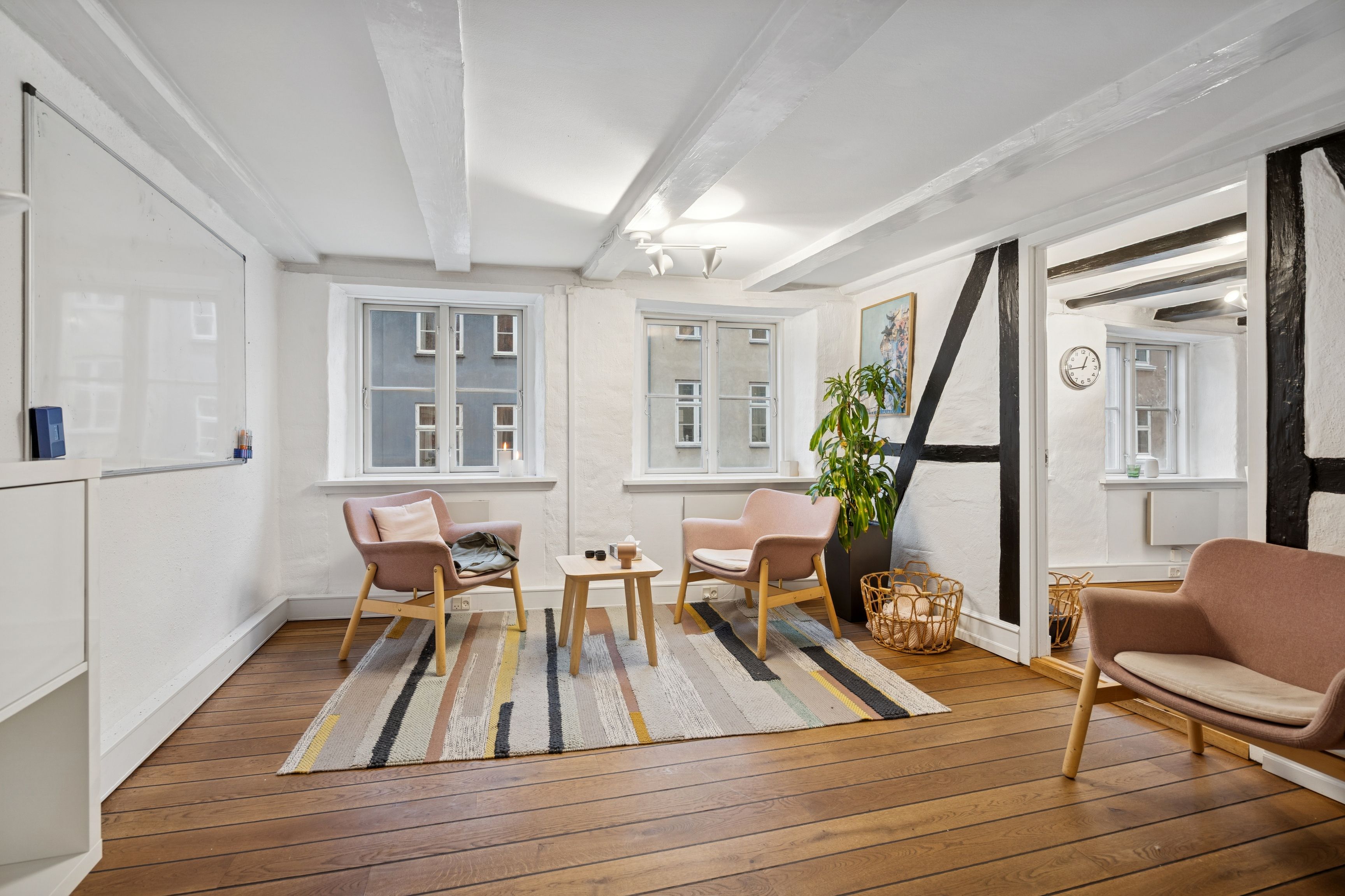 Lyst og funktionelt kontor med højloft, moderne møbler, lyse trægulve, store vinduer og en stor plante i hjørnet.