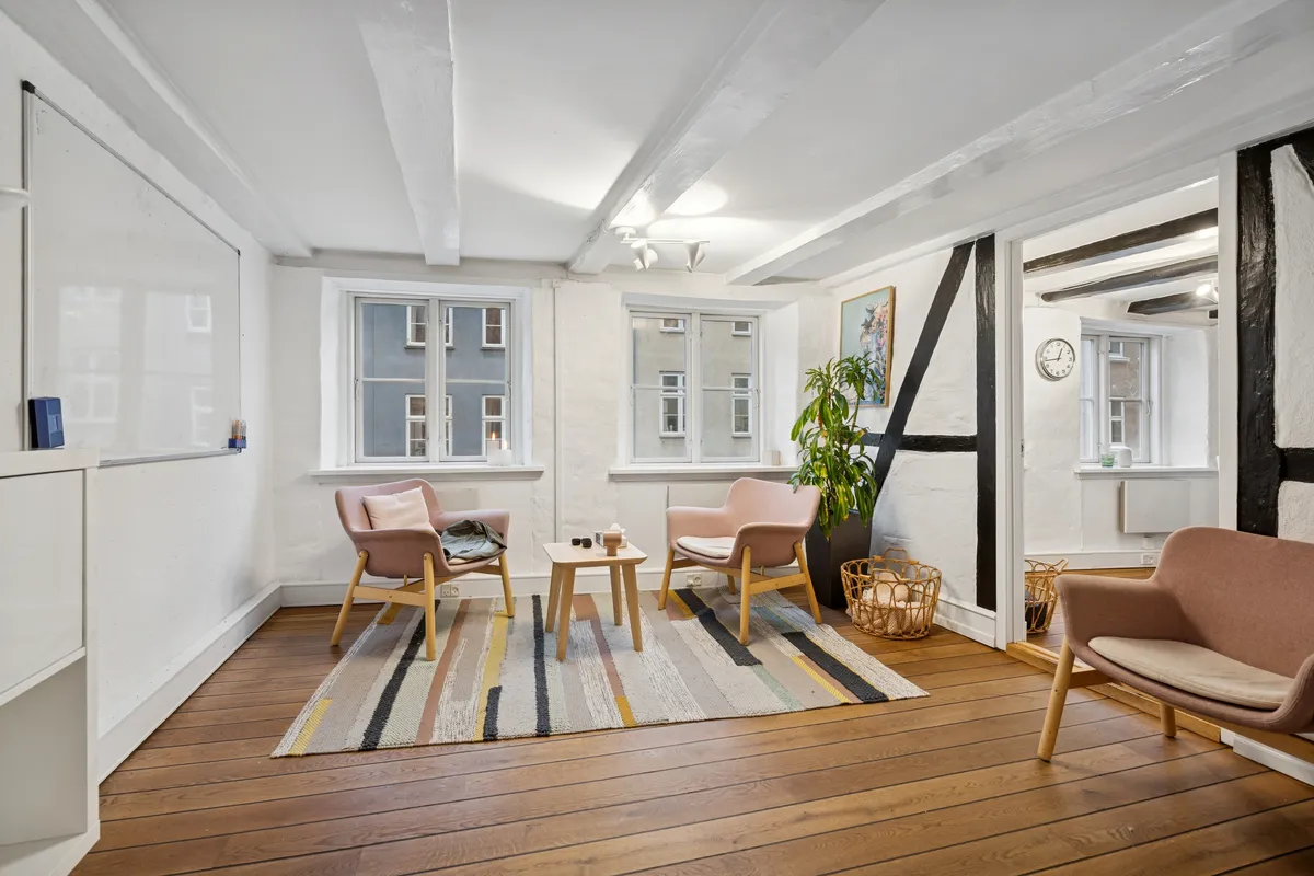 Lyst og funktionelt kontor med højloft, moderne møbler, lyse trægulve, store vinduer og en stor plante i hjørnet.